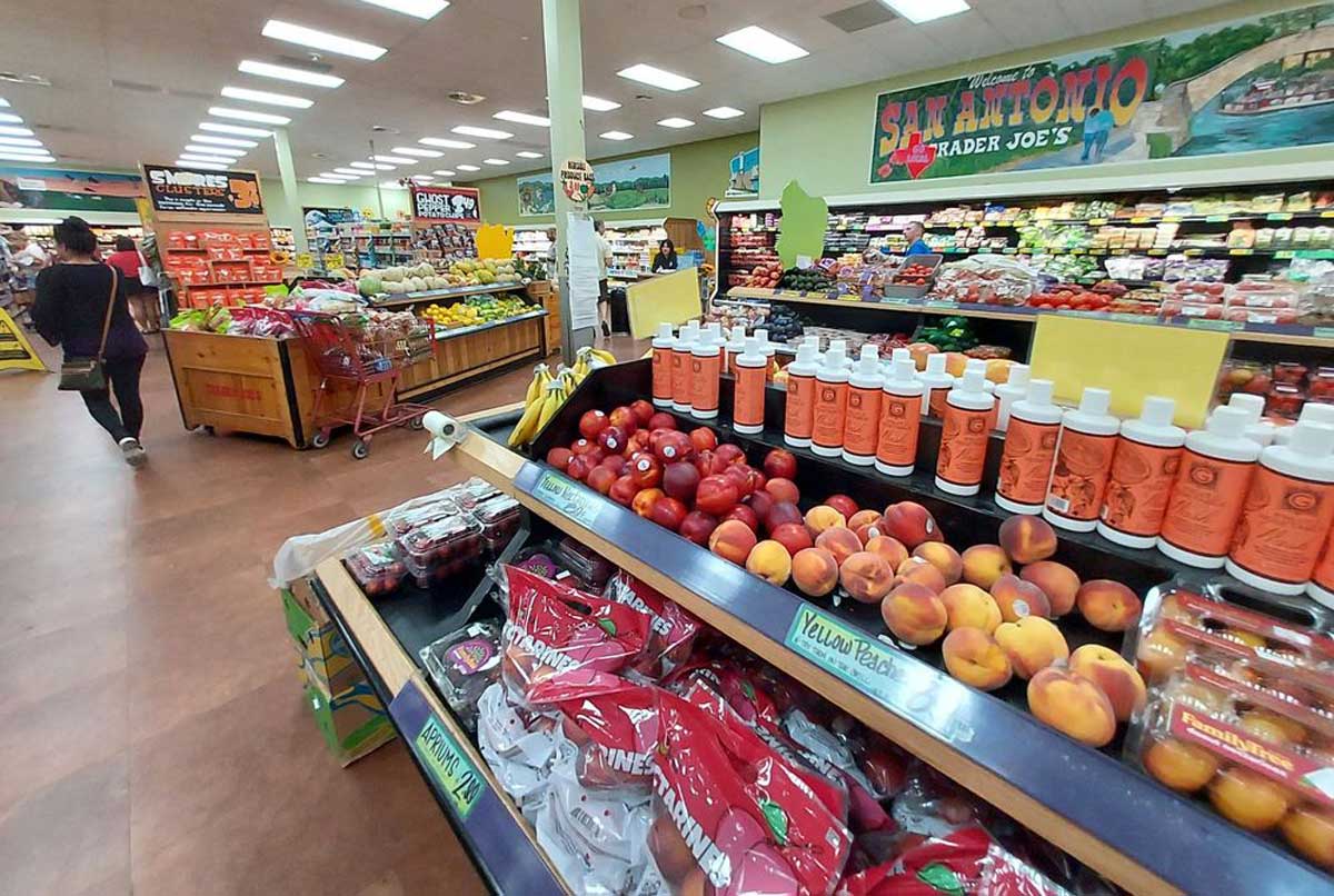 Produce section of a grocery store with peaches, apples, and bottled juice on display. A shopper walks down the aisle; colorful fruits, vegetables, and a mural reading "San Antonio" are visible in the background.