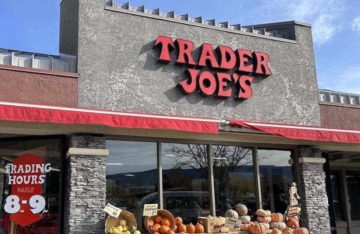 The front of a new Trader Joe's store with a large red sign, baskets of pumpkins and gourds displayed outside, and a sign showing trading hours of 8-9 on the window.