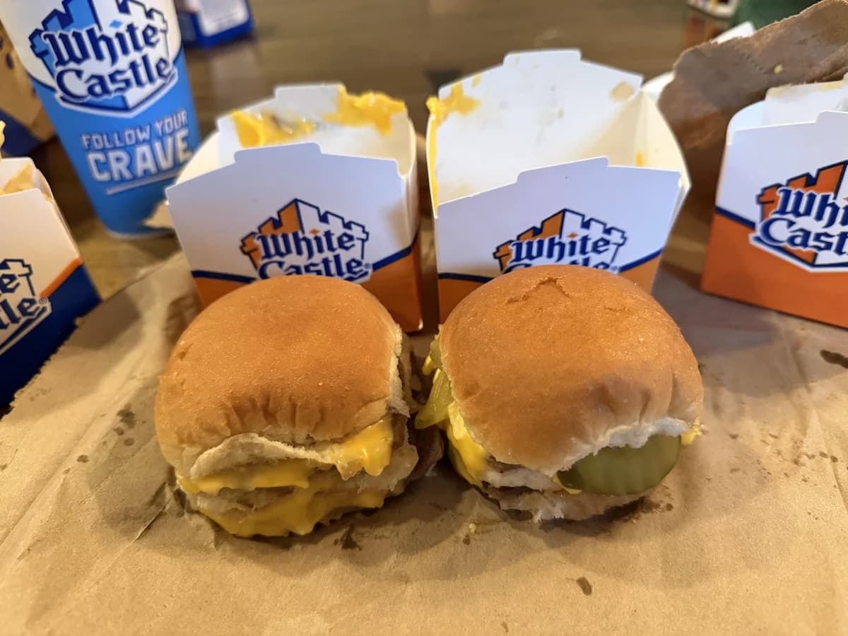 Two White Castle cheeseburgers with pickles and cheese sit on a brown paper surface, highlighting the classic look of these famously overrated fast-food burgers, with open White Castle boxes and a drink cup in the background.