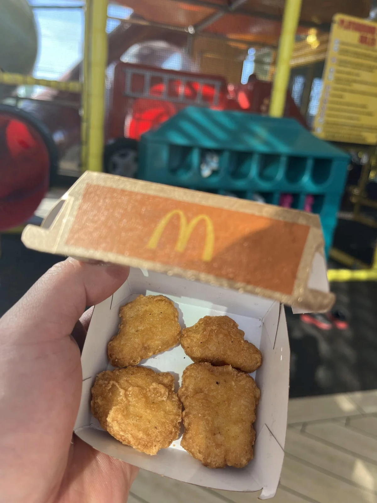 A hand holds an open McDonald's box containing four chicken nuggets, with a playground and colorful play equipment blurred in the background.