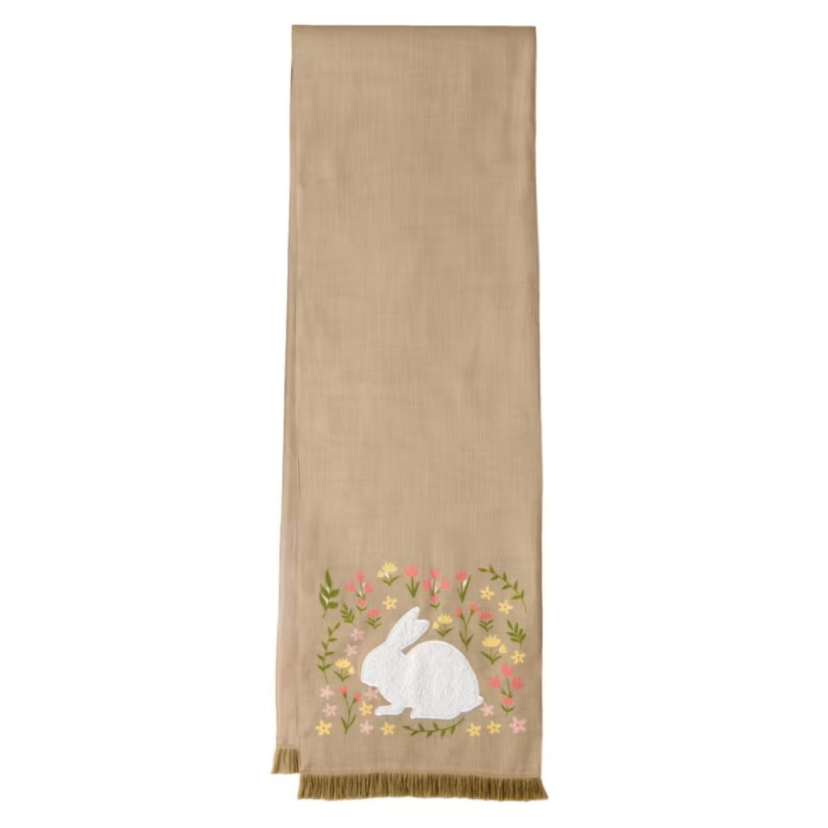 A beige table runner with fringed edges, featuring an embroidered white bunny surrounded by colorful flowers and greenery near one end.