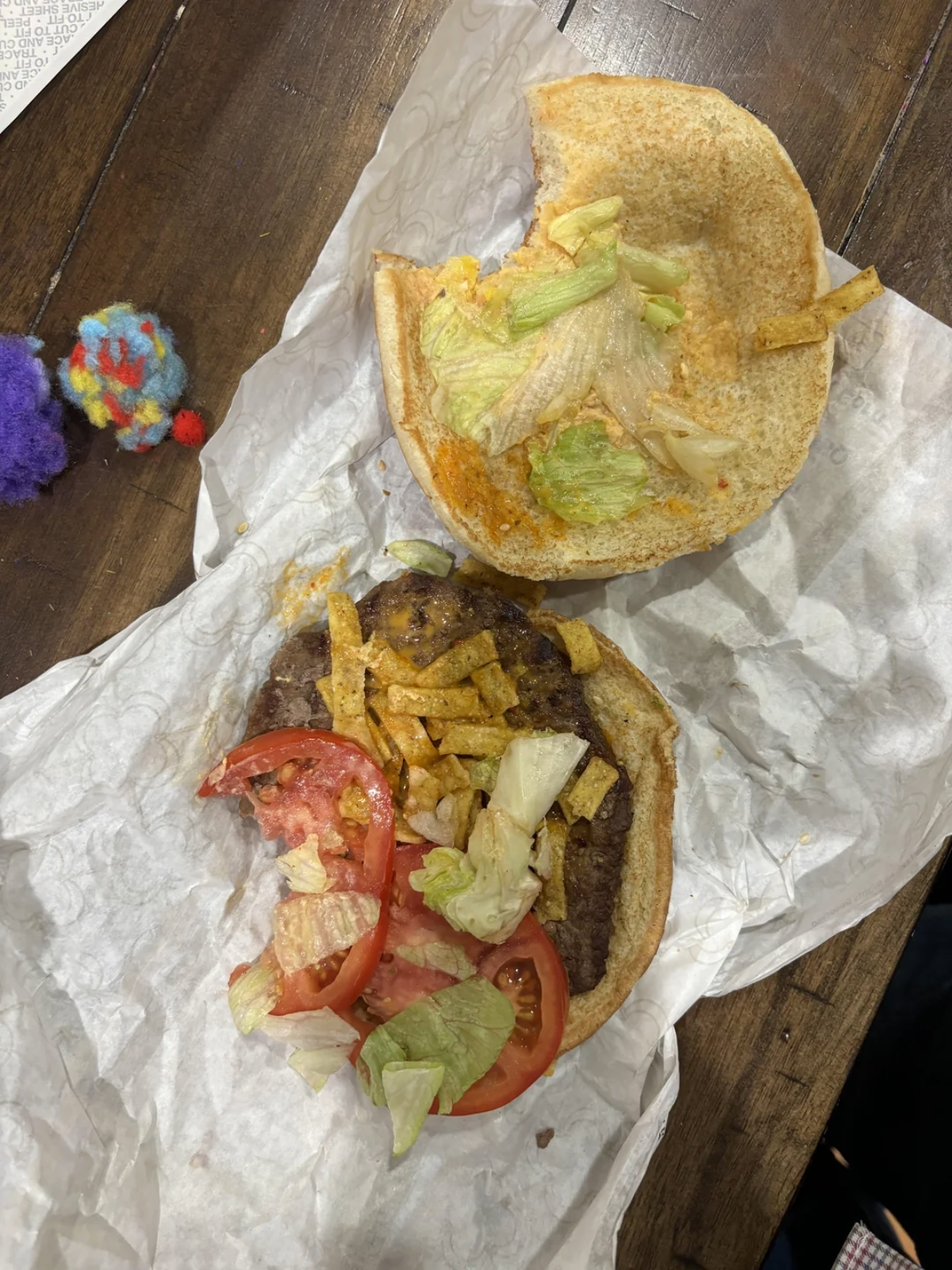 A partially assembled burger on white paper shows lettuce, tomato, tortilla strips, and a beef patty on a bun. Some lettuce and sauce are on the top bun. A colorful pom-pom toy is nearby on a wooden table.
