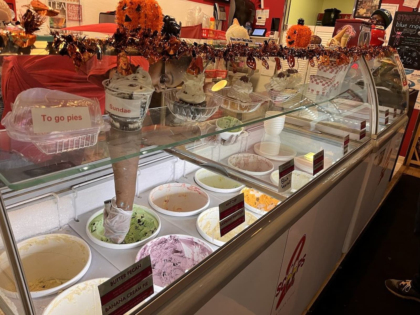 An ice cream shop display case with various ice cream tubs, cones, and toppings. Labels show flavors like butter pecan and banana cream pie. Halloween decorations with orange garland hang above the counter.