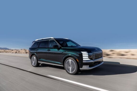 A large, dark green SUV is driving on a paved road through a desert landscape under a clear blue sky.