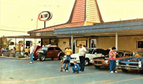 A retro photo of a 1970s family restaurant, A&W, shows a parking lot filled with classic cars, adults standing and talking, and children playing. The iconic orange A&W sign stands out on the building with its sloped roof.