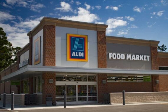 The image shows the exterior of an ALDI food market store with a large ALDI logo and "FOOD MARKET" sign above the entrance on a sunny day&mdash;perfect for shopping St. Patrick's Day items at Aldi. The building features brick, glass doors, and large windows.