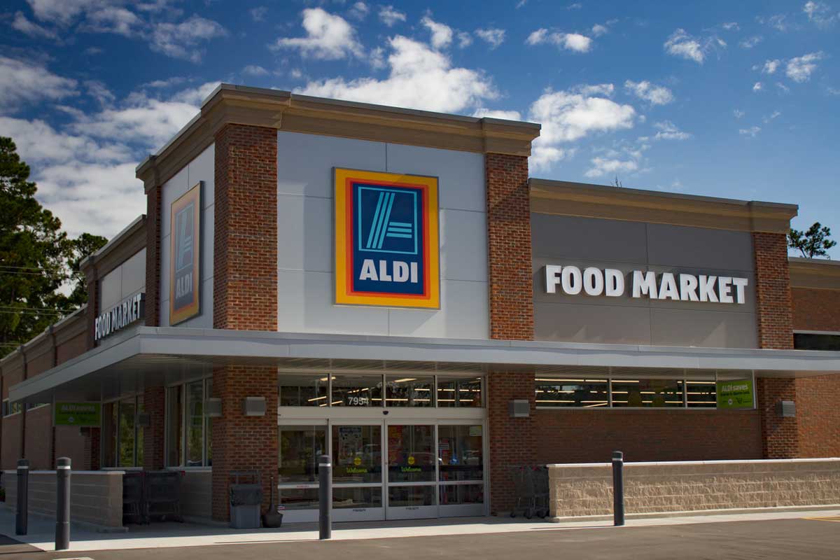 The image shows the exterior of an ALDI food market store with a large ALDI logo and "FOOD MARKET" sign above the entrance on a sunny day—perfect for shopping St. Patrick's Day items at Aldi. The building features brick, glass doors, and large windows.