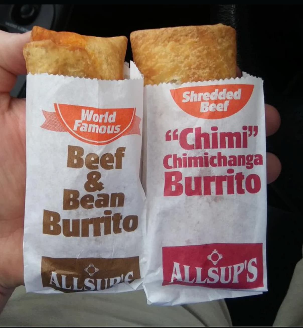 A hand holds two Allsup's burritos: one in a wrapper labeled "World Famous Beef & Bean Burrito" and the other in a wrapper labeled "Shredded Beef 'Chimi' Chimichanga Burrito.