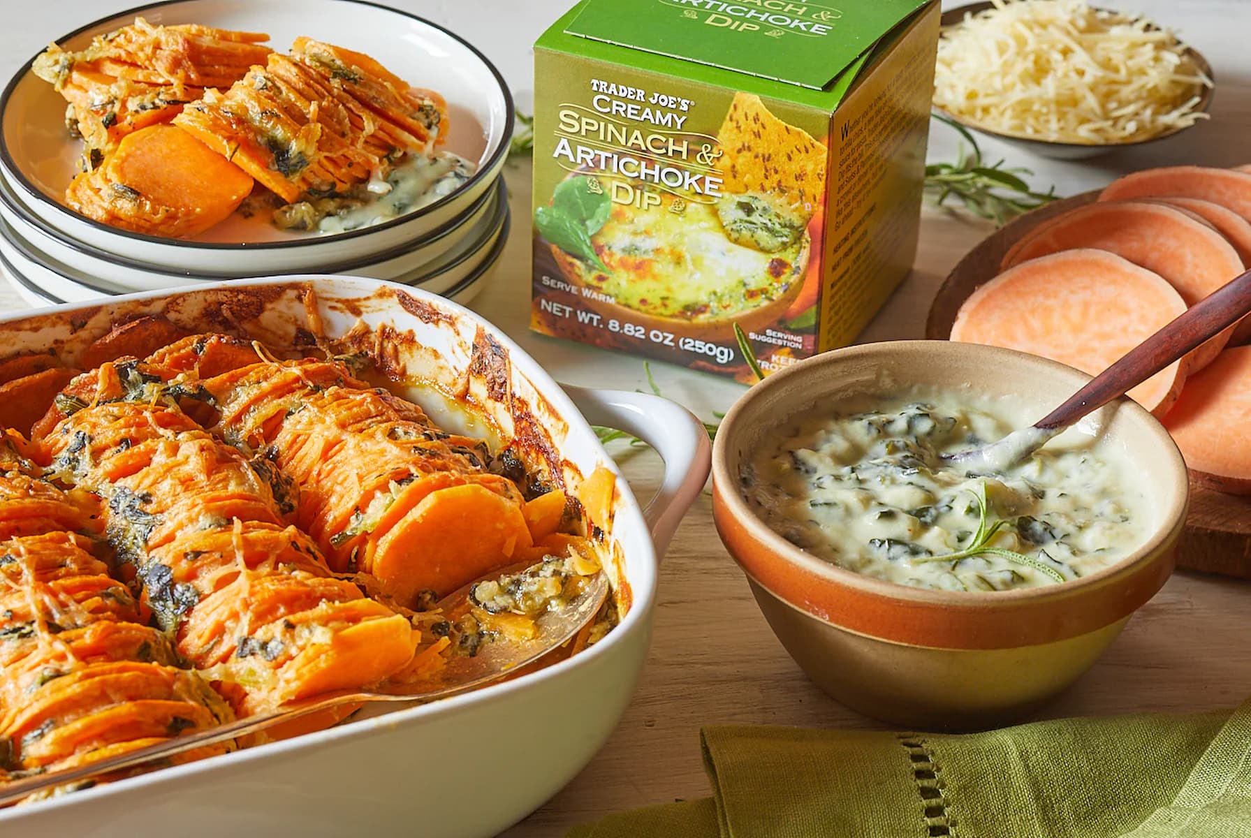 A baked sweet potato and spinach artichoke dip casserole sits in a white dish, with bowls of dip, a box of Trader Joe’s Spinach & Artichoke Dip, shredded cheese, sweet potato slices, and a green napkin nearby.
