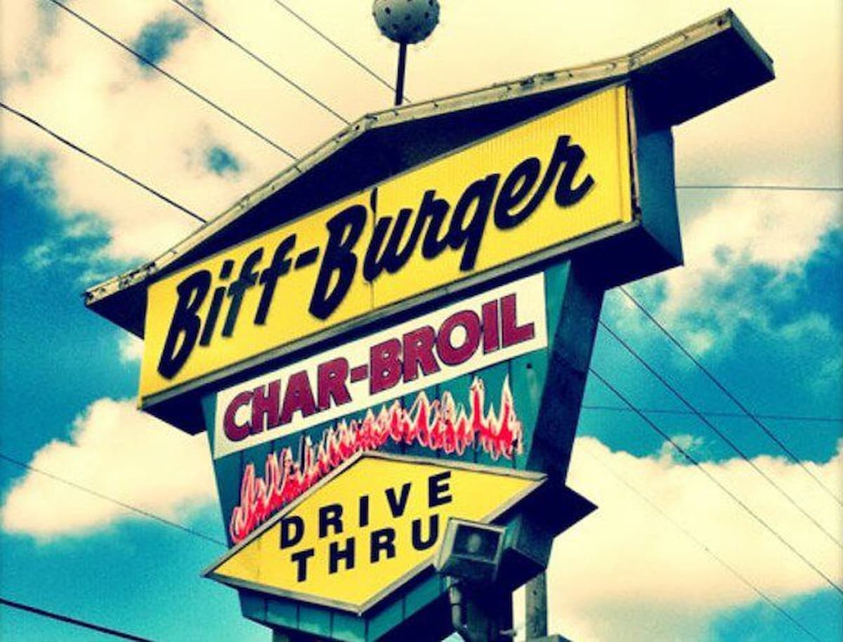 A retro yellow and black sign reads "Biff-Burger" with a smaller sign below saying "Char-Broil Drive Thru" against a blue sky with clouds and power lines.