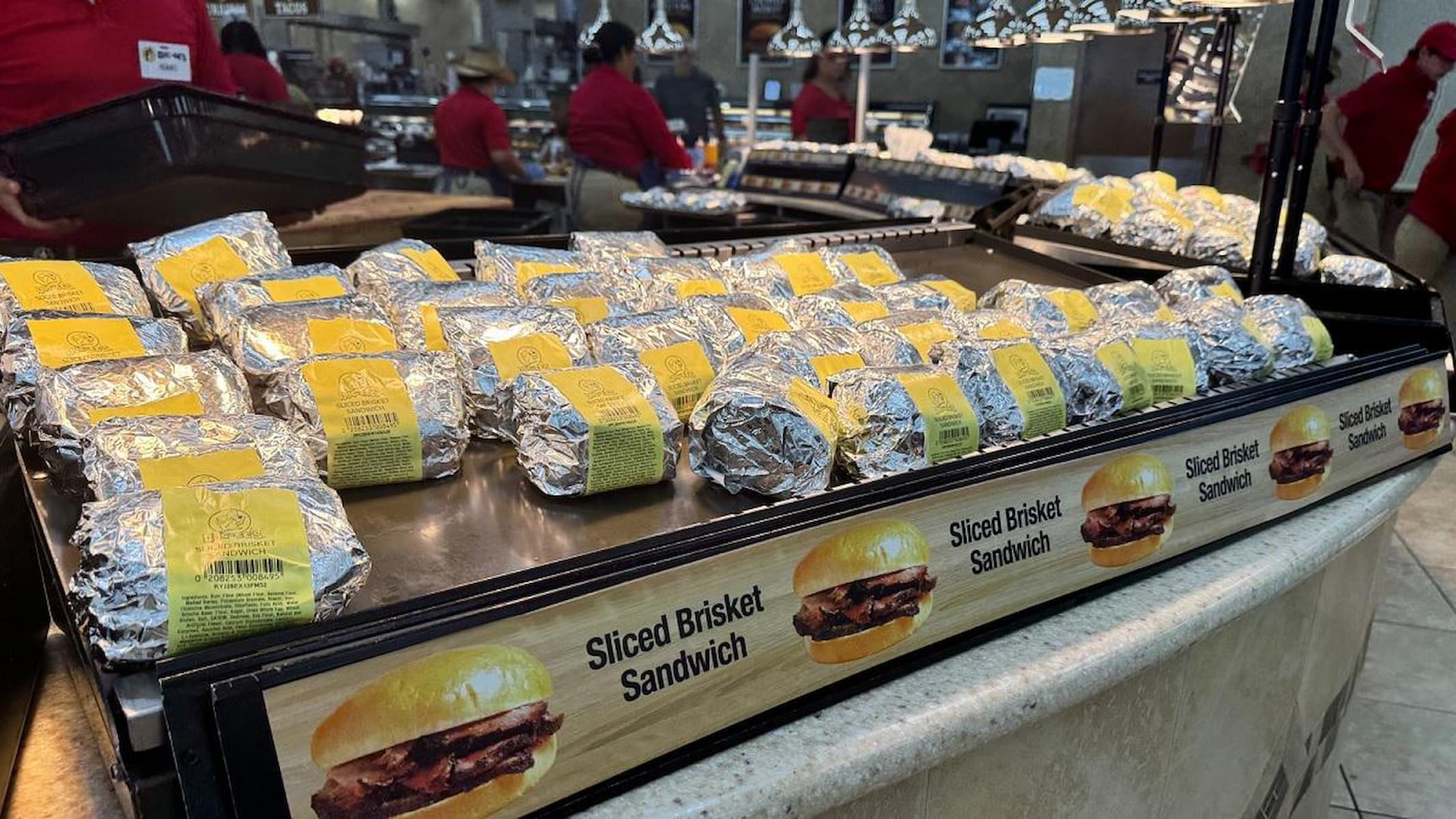 Rows of foil-wrapped sandwiches labeled "Sliced Brisket Sandwich" are displayed on a counter at a deli or market, with employees in red shirts working in the background.
