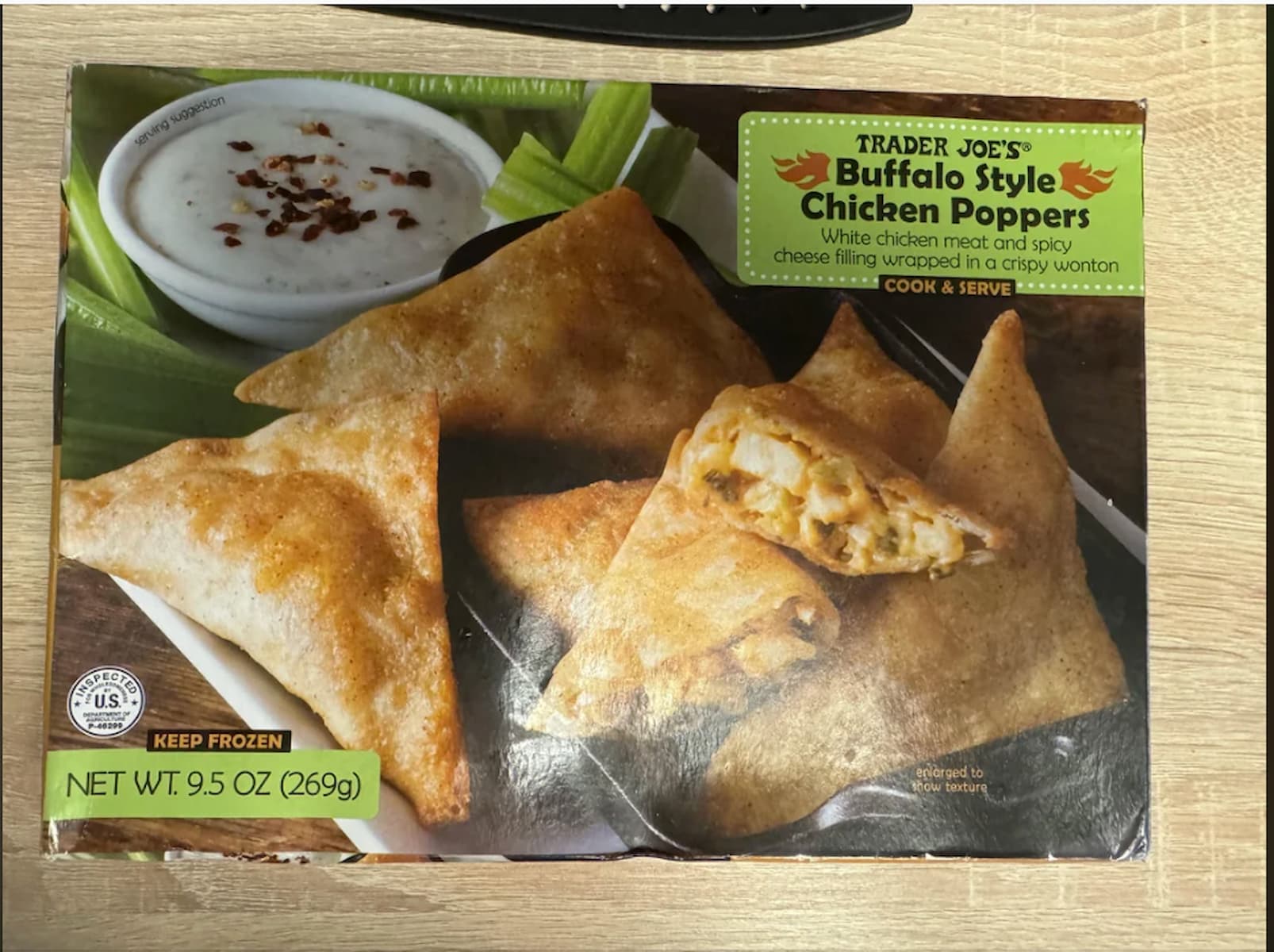 Box of Trader Joe’s Buffalo Style Chicken Poppers, featuring crispy wonton triangles filled with chicken and cheese, with a side of ranch dipping sauce. The box is on a light wood surface.
