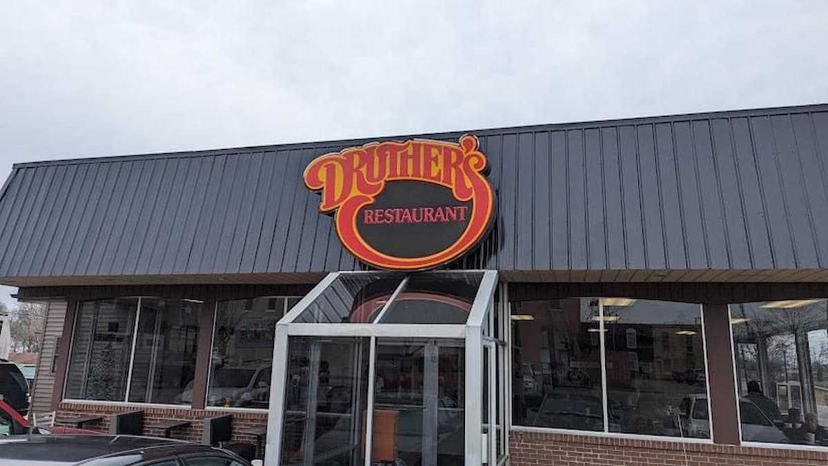 Exterior view of Druther's Restaurant with a red and yellow sign above the entrance. The building has large windows and a glass vestibule at the front. The sky is overcast.