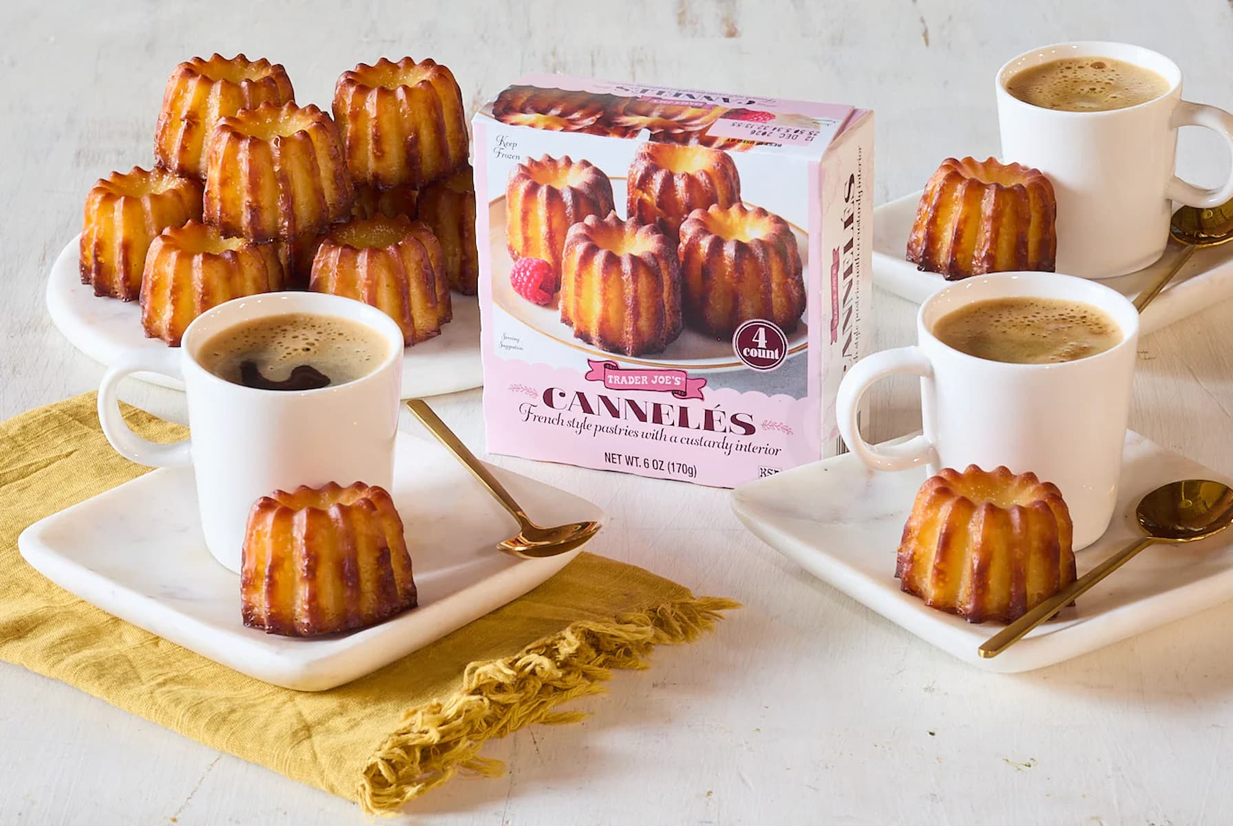 Three white cups of coffee on square plates, each with a cannelé pastry, sit on a table with more cannelés stacked nearby. A Trader Joe’s Cannelés box is in the center. Gold spoons and a yellow napkin complete the setting.