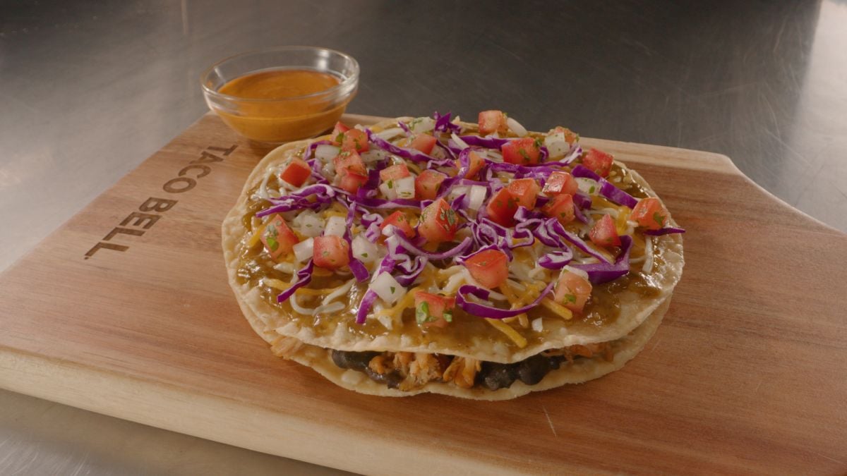 A Taco Bell Mexican Pizza with layers of tortillas, beans, cheese, shredded purple cabbage, diced tomatoes, and sauce sits on a wooden board, with a small bowl of sauce on the side.