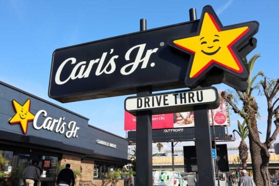 A Carl&rsquo;s Jr. fast food restaurant with a large sign featuring a yellow smiling star logo and the words &ldquo;Carl&rsquo;s Jr.&rdquo; and &ldquo;Drive Thru&rdquo; stands in front of the building, undisturbed by bankruptcy rumors. Palm trees and a clear blue sky are visible.