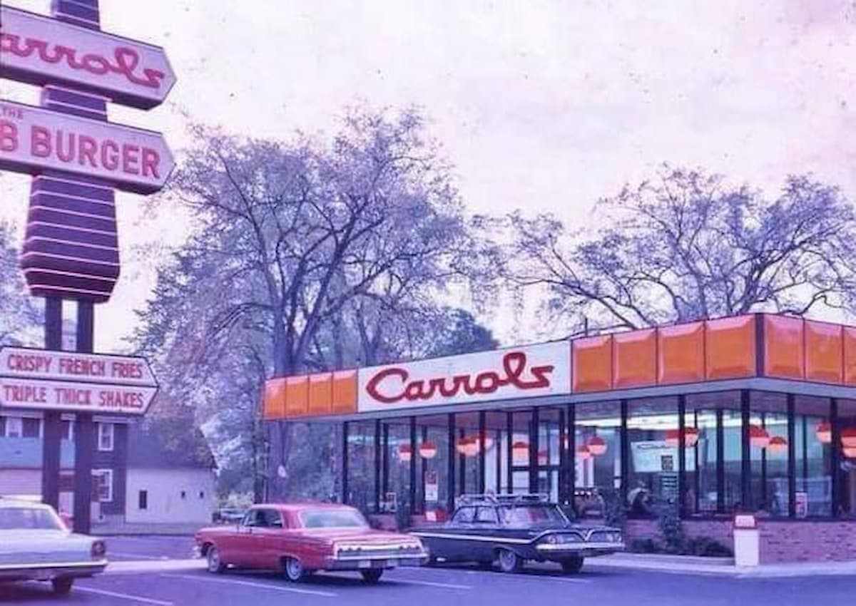 A vintage photo of a Carrols fast food restaurant with a retro sign, classic cars parked outside, and trees in the background. The restaurant features large windows and orange accents.