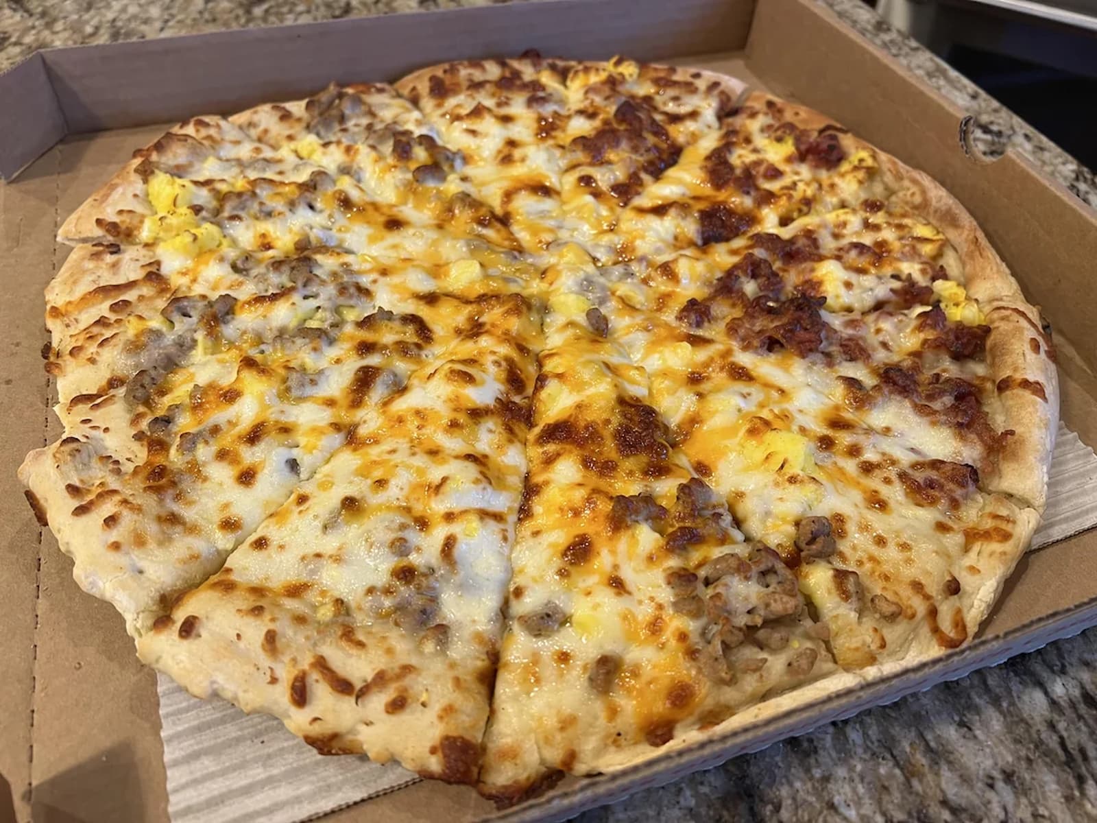 A pizza sliced into ten pieces sits in an open cardboard box. The pizza has a golden, bubbly cheese topping with spots of sausage and what appears to be scrambled eggs. The crust is thin and lightly browned.