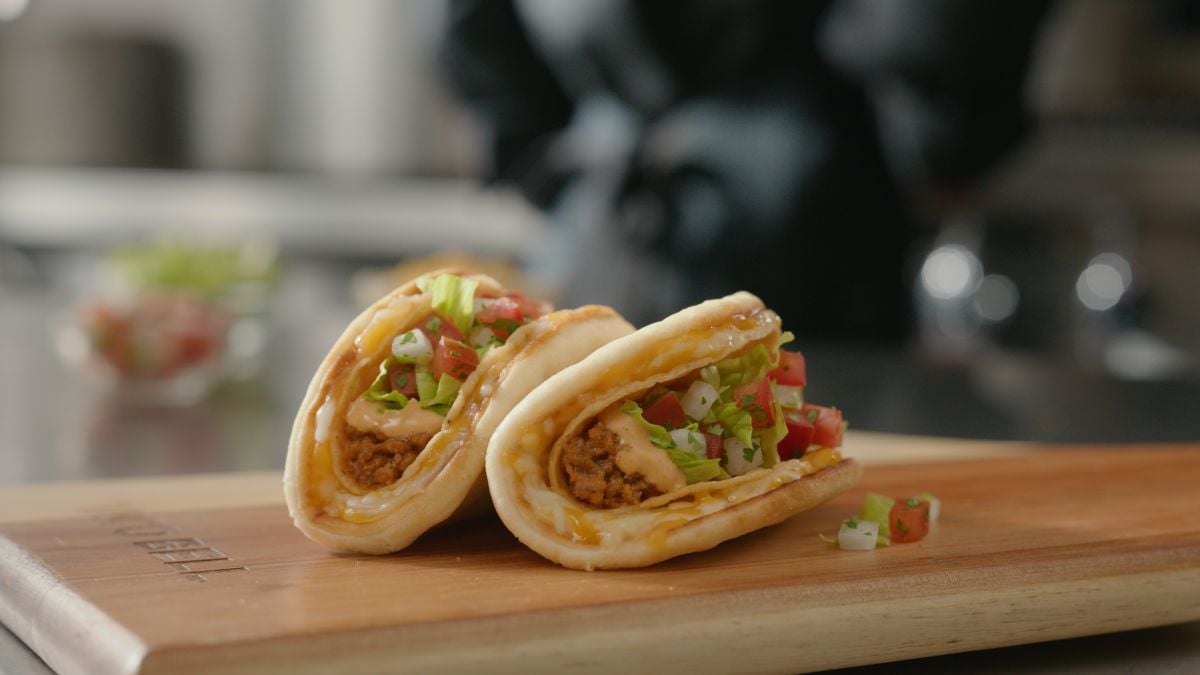 Two folded taco sandwiches filled with crispy seasoned meat, lettuce, diced tomatoes, and cheese sit on a wooden board. The background is blurred, focusing on the fresh ingredients and soft flatbread.