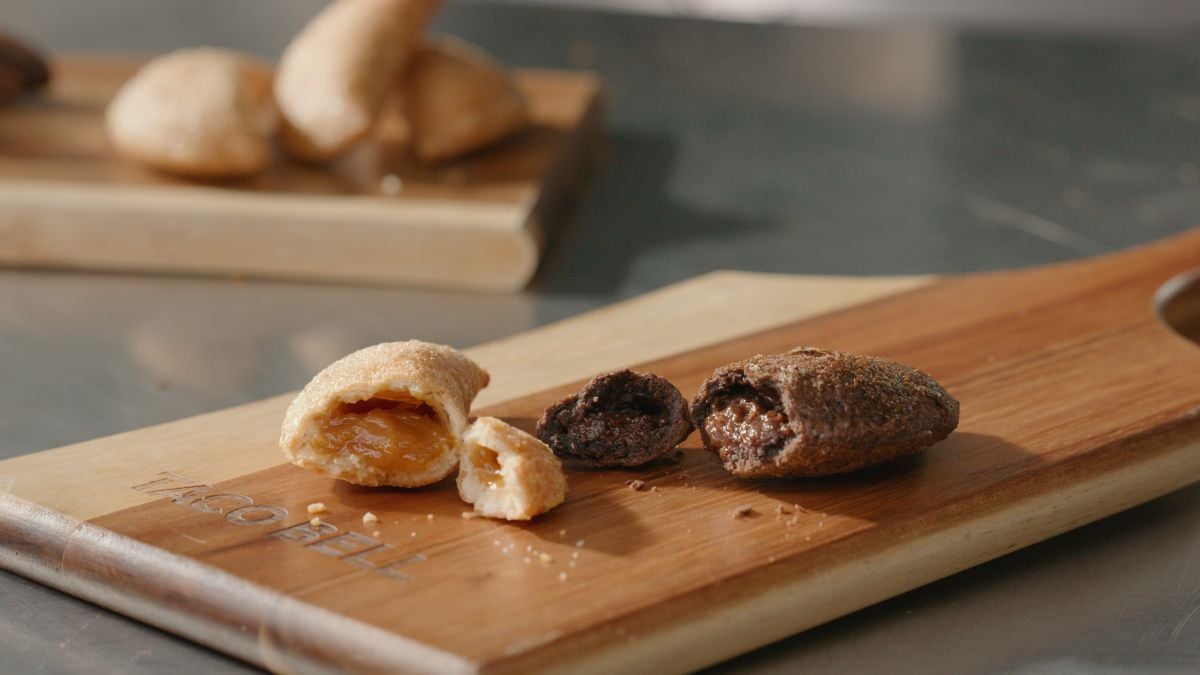 A close-up of a wooden serving board with two pastries, one with a golden brown crust and fruit filling and another with a dark crust and chocolate filling, both broken open. More pastries are blurred in the background.