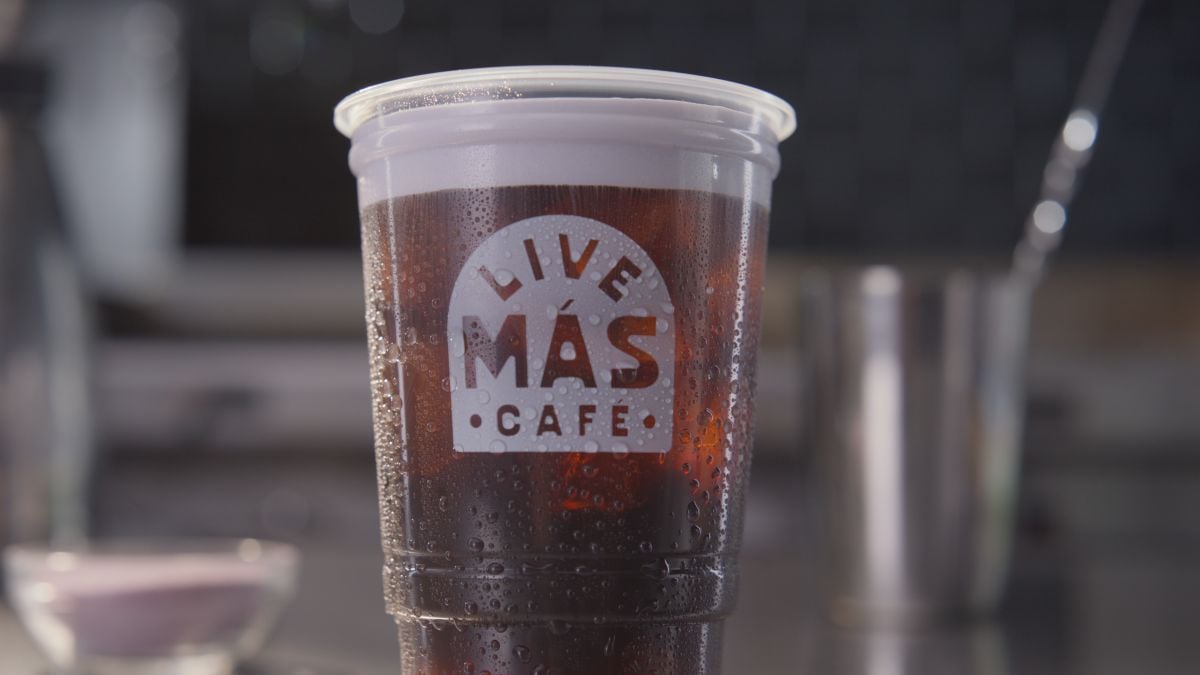 A plastic cup filled with a dark, cold beverage, covered in condensation. The cup has the "Live Más Café" logo printed on it. The background is blurred, with some kitchen items faintly visible.