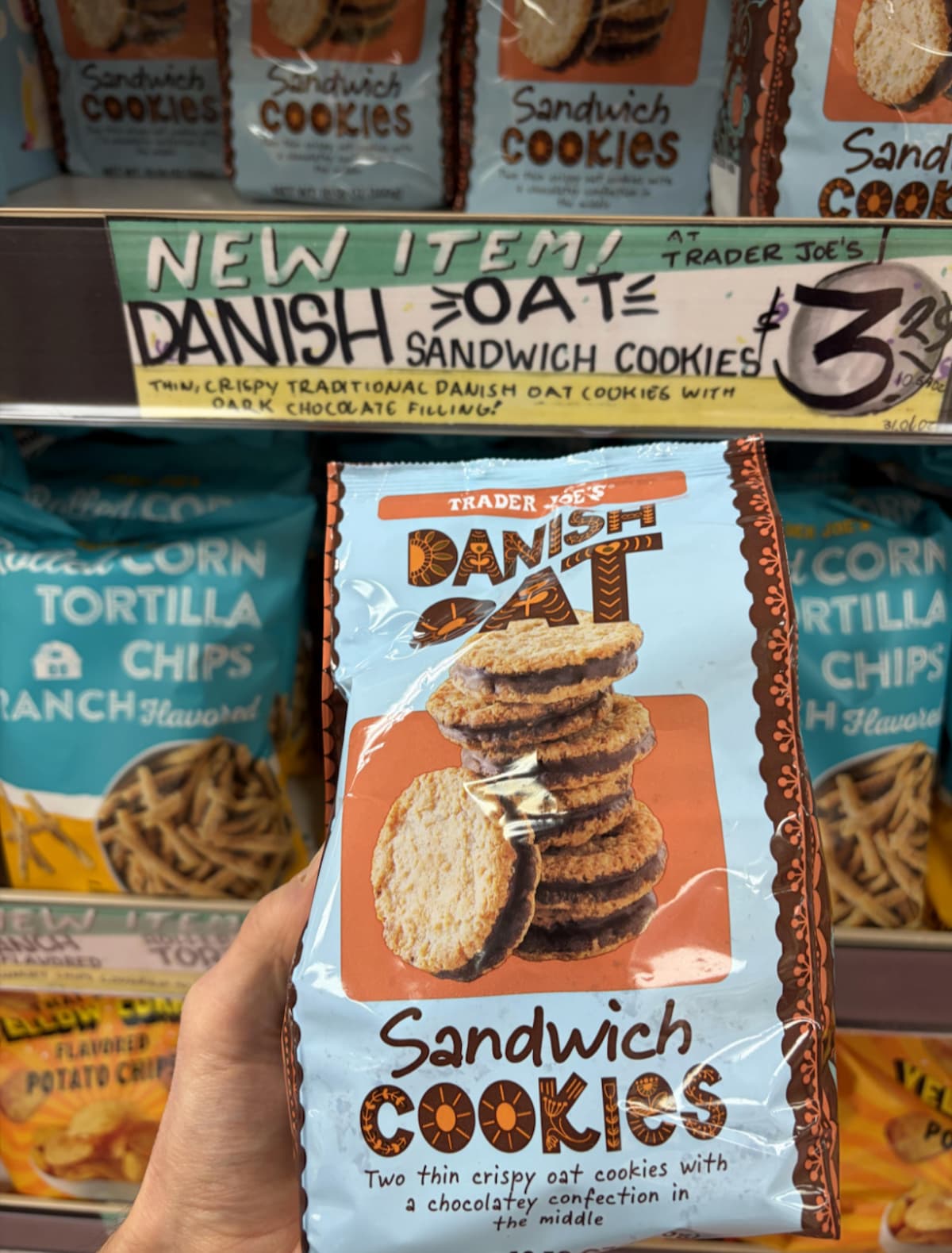 A hand holds a bag of Trader Joe’s Danish Oat Sandwich Cookies in front of a store shelf. The blue bag shows images of the cookies, and a sign above advertises them as a new item for $3.99.