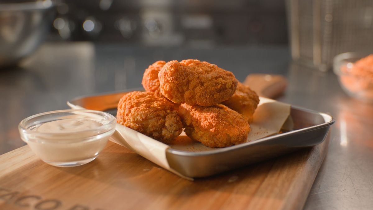 Four crispy, breaded chicken wings sit on parchment paper in a metal tray next to a small glass bowl of dipping sauce, all placed on a wooden board in a kitchen setting.