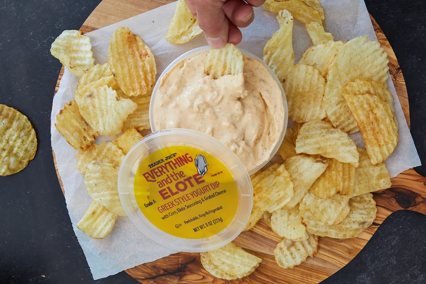 A hand dips a ridged potato chip into a container of Everything and the Elote Greek Style Yogurt Dip, surrounded by more potato chips on a wooden board lined with parchment paper.