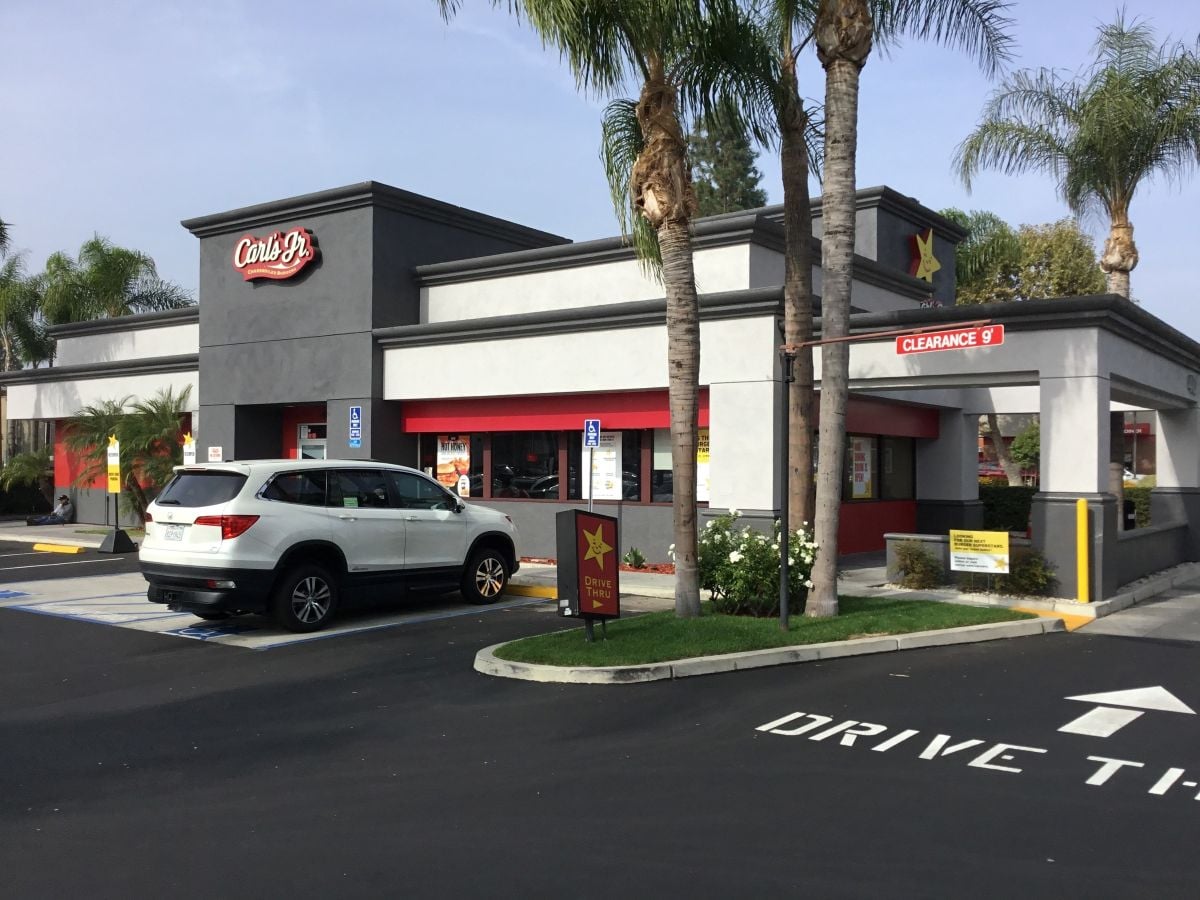 A white SUV is parked outside a Carl’s Jr. fast food restaurant with palm trees around the building. A drive-thru sign and menu board are visible near the entrance of the location, once rumored to face bankruptcy.