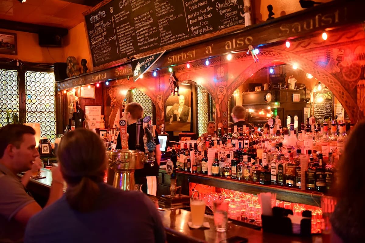 A cozy, dimly lit bar with colorful string lights, shelves full of liquor bottles, and a bartender serving drinks to customers seated at the counter. A chalkboard menu and framed pictures decorate the walls.