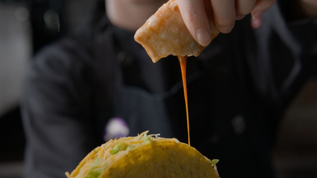 A hand dips a crispy taco shell filled with lettuce and shredded cheese into a cup of orange sauce, with the sauce dripping onto the taco. The background is blurred, focusing on the food.