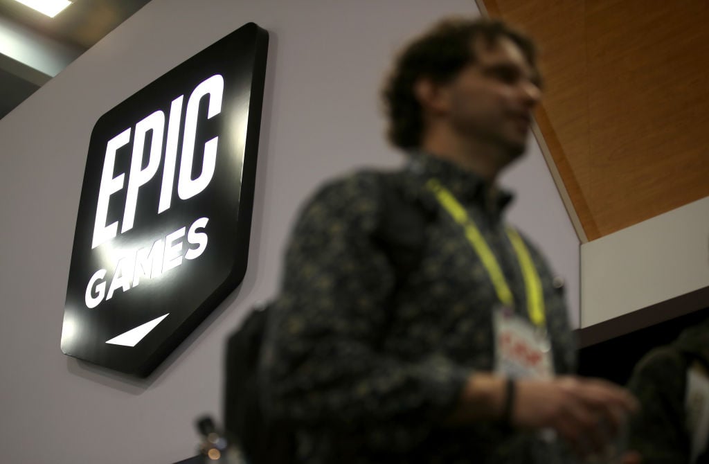 A large illuminated Epic Games sign on a wall, with a person in the foreground standing out of focus wearing a lanyard and backpack—an image set against the backdrop of the 2026 layoffs.