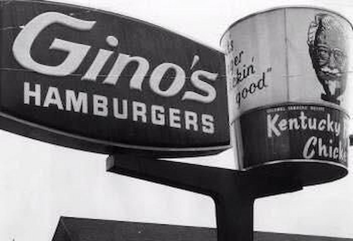 Black and white photo of two signs: one for "Gino’s Hamburgers" and another shaped like a large Kentucky Fried Chicken bucket with Colonel Sanders’ image and the slogan "It’s finger lickin’ good.