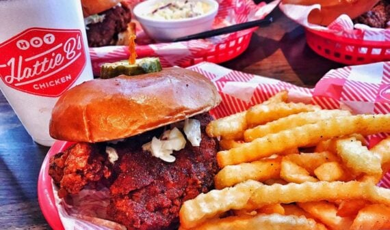 A basket of crinkle-cut fries and a spicy fried chicken sandwich with pickles sits on red-and-white checkered paper next to a Hattie B&rsquo;s cup. Other baskets of food are visible in the background.
