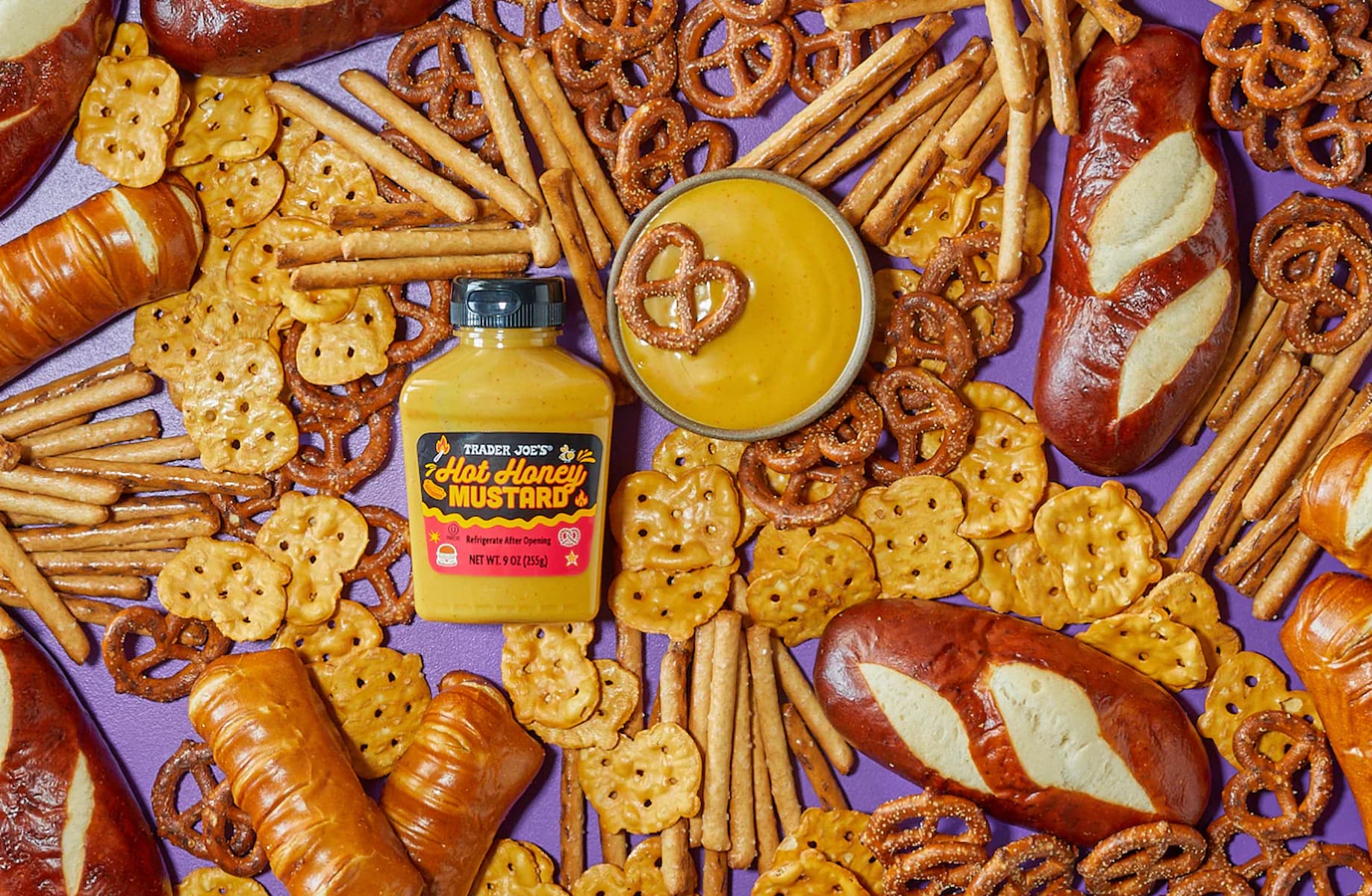 A spread of pretzel rolls, pretzel twists, pretzel sticks, and mini pretzel crackers surrounds a jar of Trader Joe’s Bold Honey Mustard and a bowl of mustard dip on a purple background.