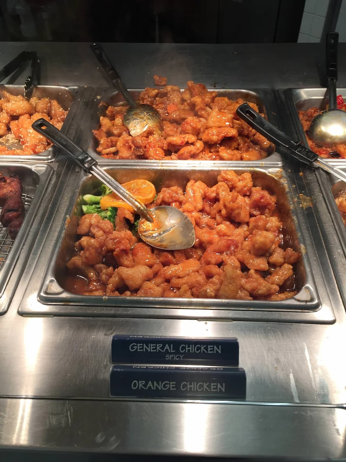 A buffet tray filled with orange chicken, featuring pieces of crispy chicken in an orange sauce, with a serving spoon resting inside. The label below reads "ORANGE CHICKEN.