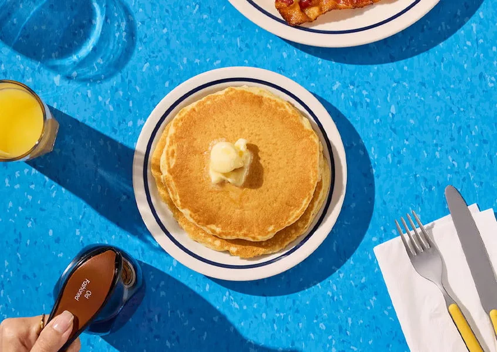 A plate with two pancakes topped with butter sits on a blue table, next to a glass of orange juice, a syrup bottle, and a plate with bacon. A napkin with a fork and knife is also visible.