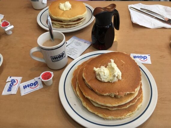 A plate of three pancakes with a dollop of butter sits next to a mug of coffee, syrup pitcher, and IHOP-branded creamers and sugar packets on a wooden table&mdash;perfect for enjoying your Free Pancakes offer. Another stack waits in the background.