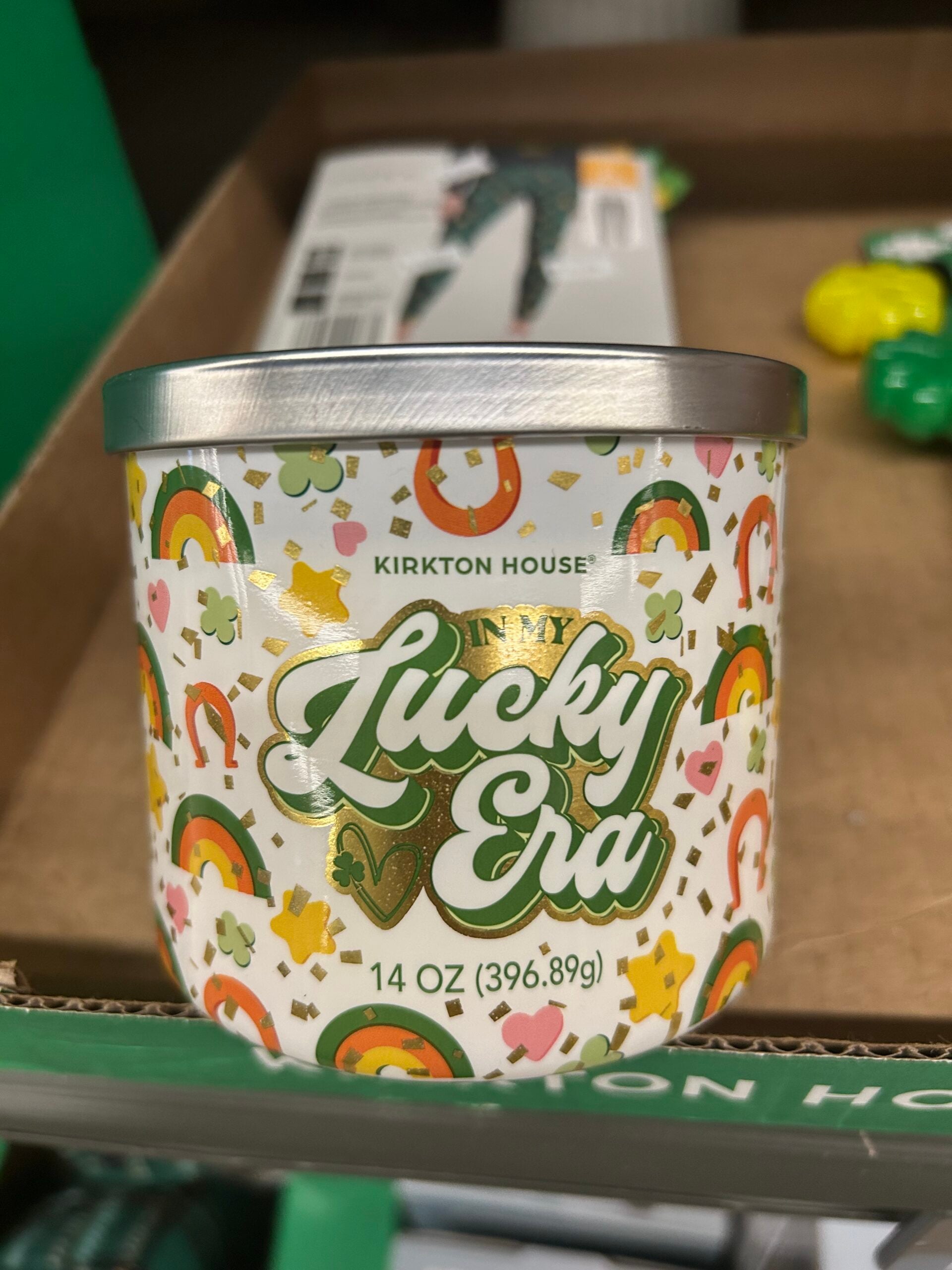 A candle with a festive design featuring rainbows, stars, and shamrocks. The label reads "Kirkton House Lucky Era" and shows the weight as 14 oz (396.89g). The candle is displayed on a store shelf.