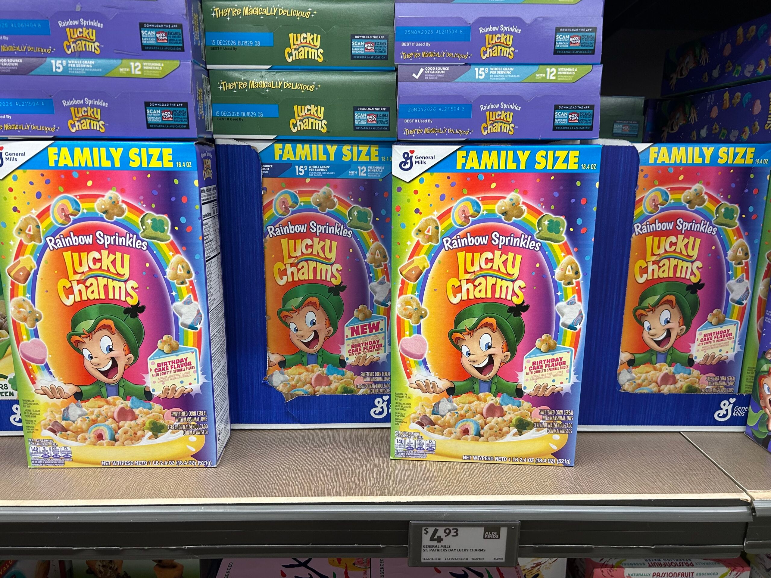 Family size boxes of Rainbow Sprinkles Lucky Charms cereal are displayed on a store shelf. The boxes feature a cartoon leprechaun and colorful marshmallow shapes. Price tag reads £4.93.