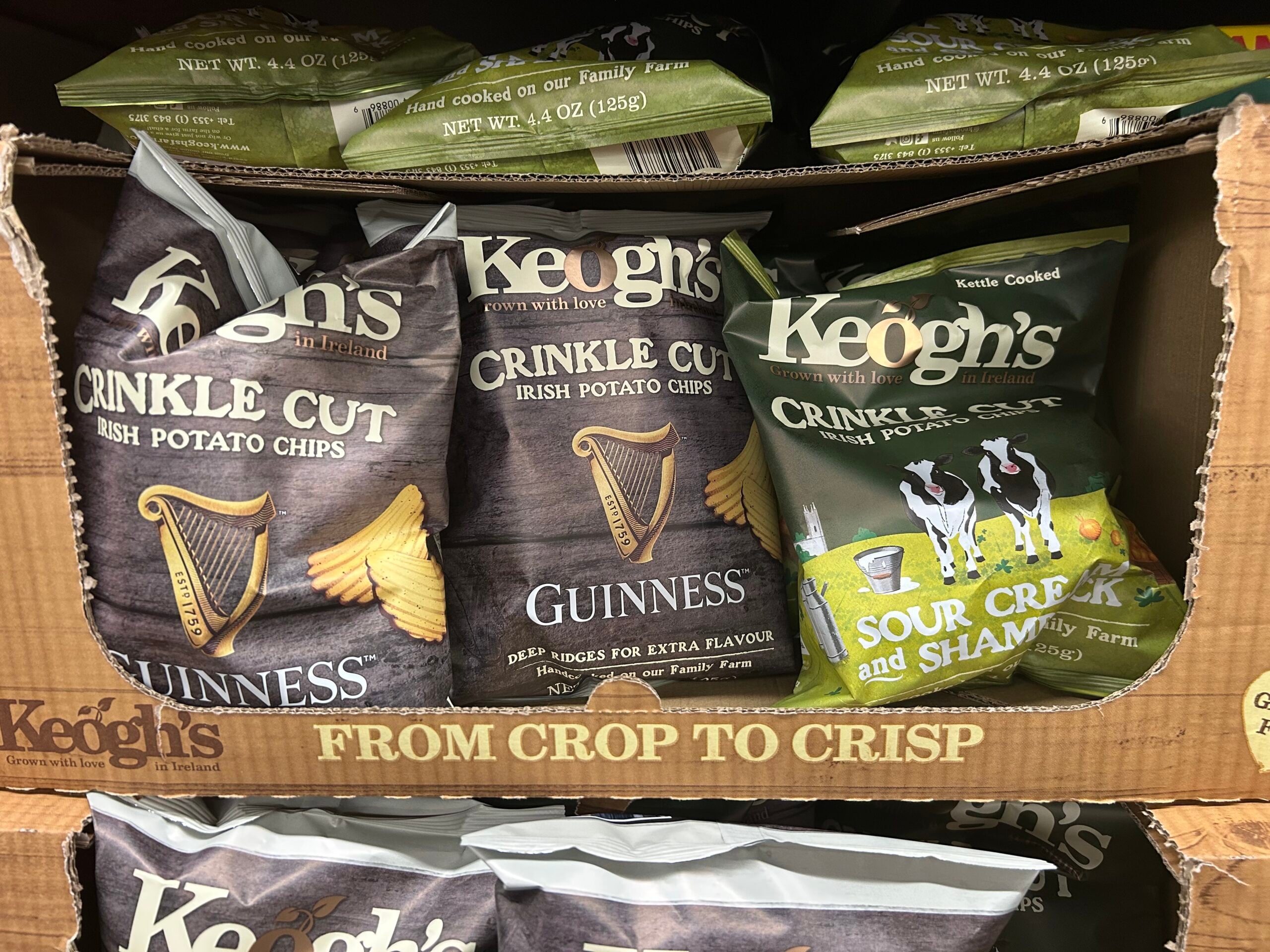 Bags of Keogh’s Irish potato chips on a store shelf, featuring two flavors: Guinness Crinkle Cut and Sour Cream & Shamrock. The packaging highlights Irish themes and imagery.