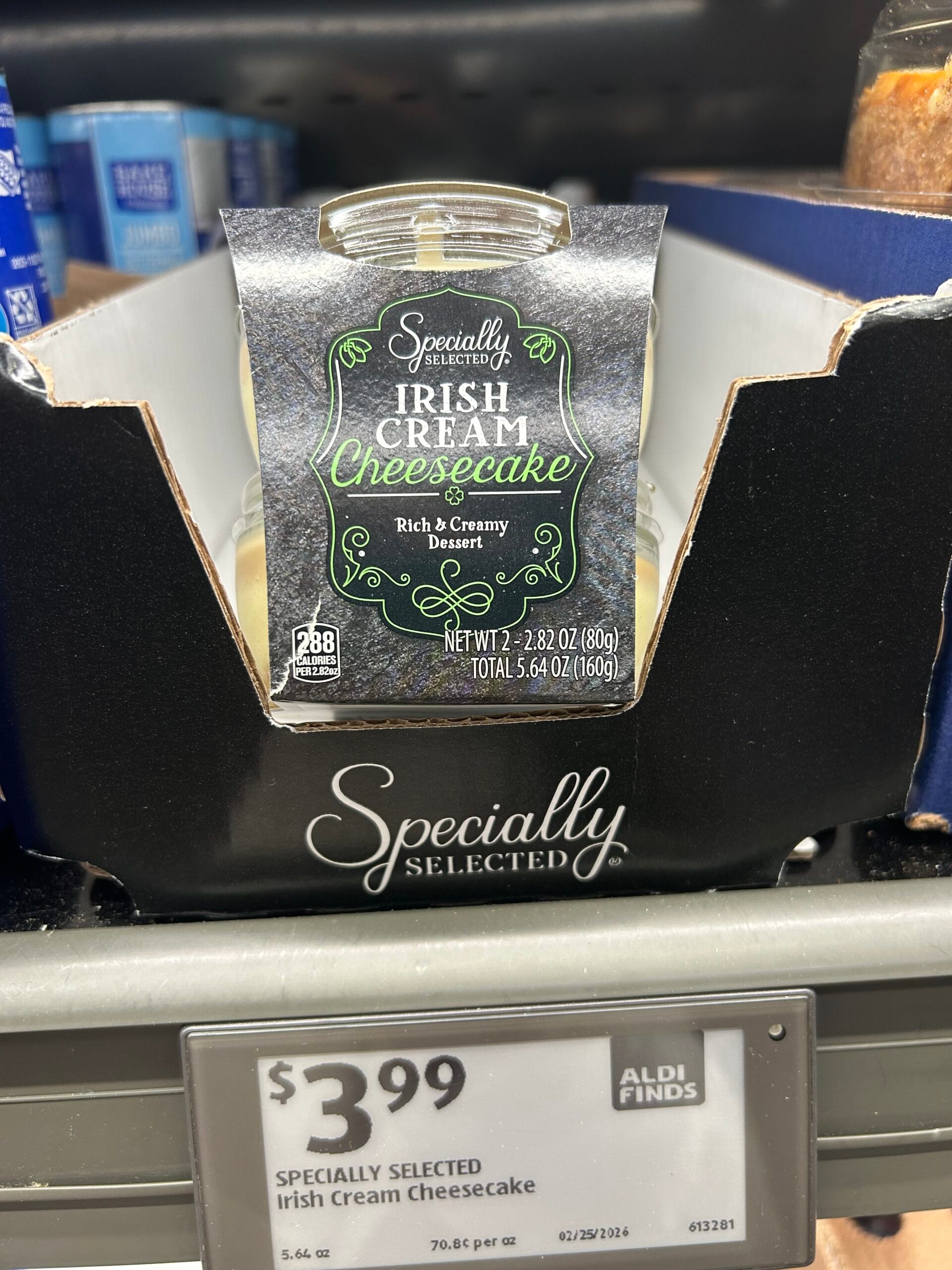 A glass jar of Specially Selected Irish Cream Cheesecake dessert is displayed in a black box on a store shelf, priced at $3.99 at ALDI. The label highlights "Rich & Creamy Dessert.