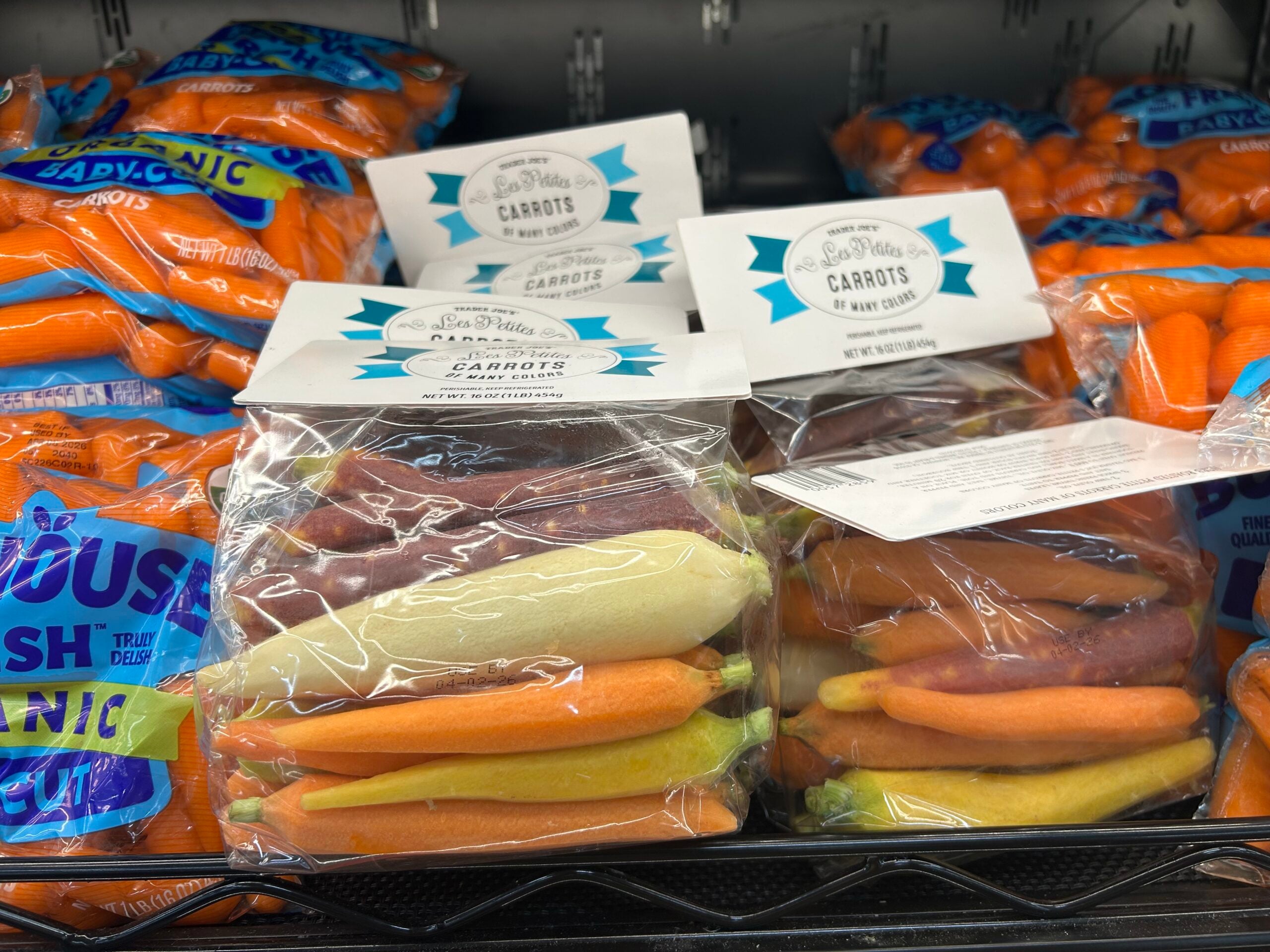 Bags of orange baby carrots sit on shelves, with clear bags of multicolored carrots—yellow, purple, and orange—in the foreground, all displayed in a grocery store produce section.