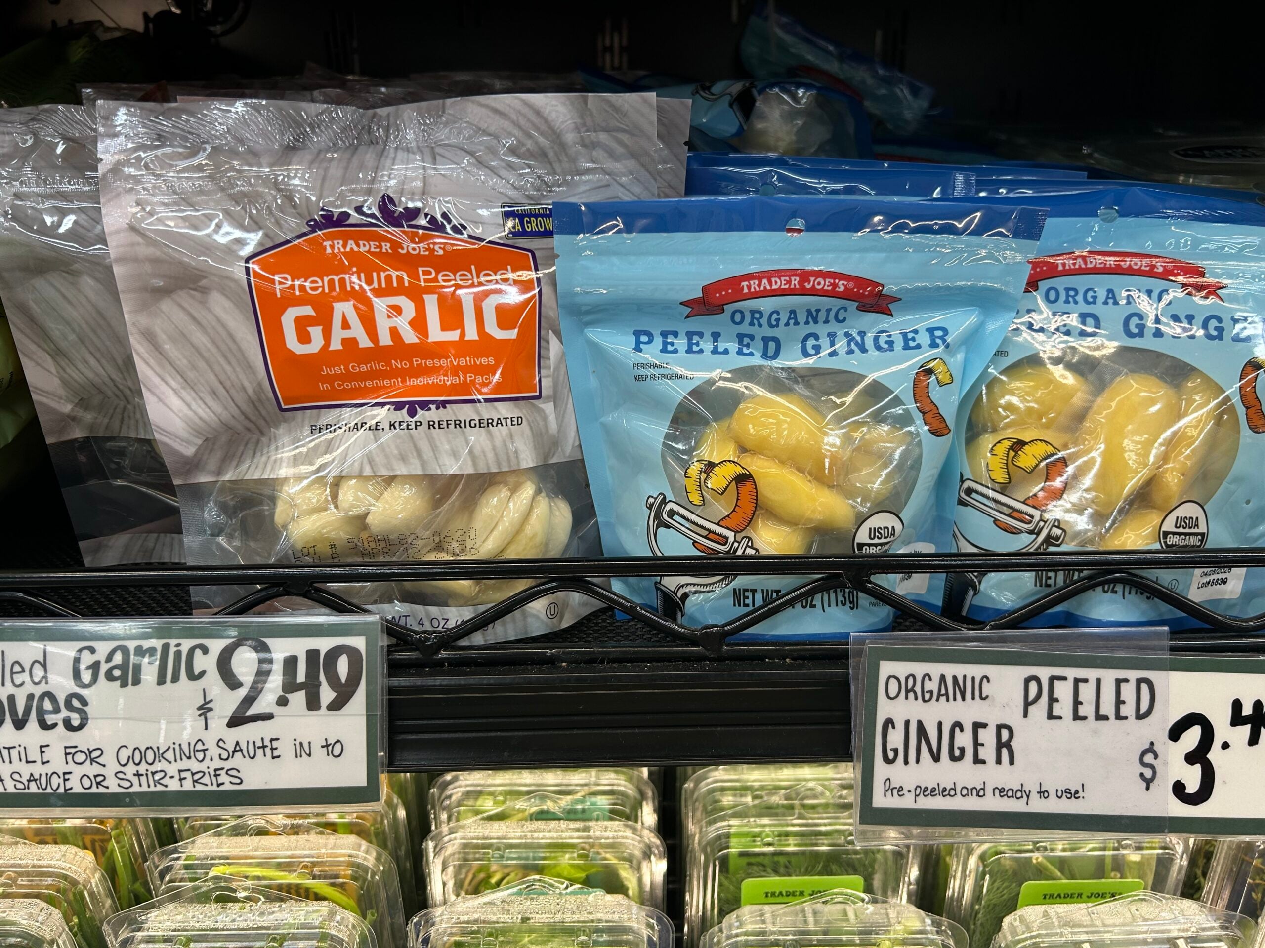 Bags of Trader Joe’s Premium Peeled Garlic and Organic Peeled Ginger are displayed on a refrigerated shelf, with price tags showing $2.49 for garlic and $3.49 for ginger.