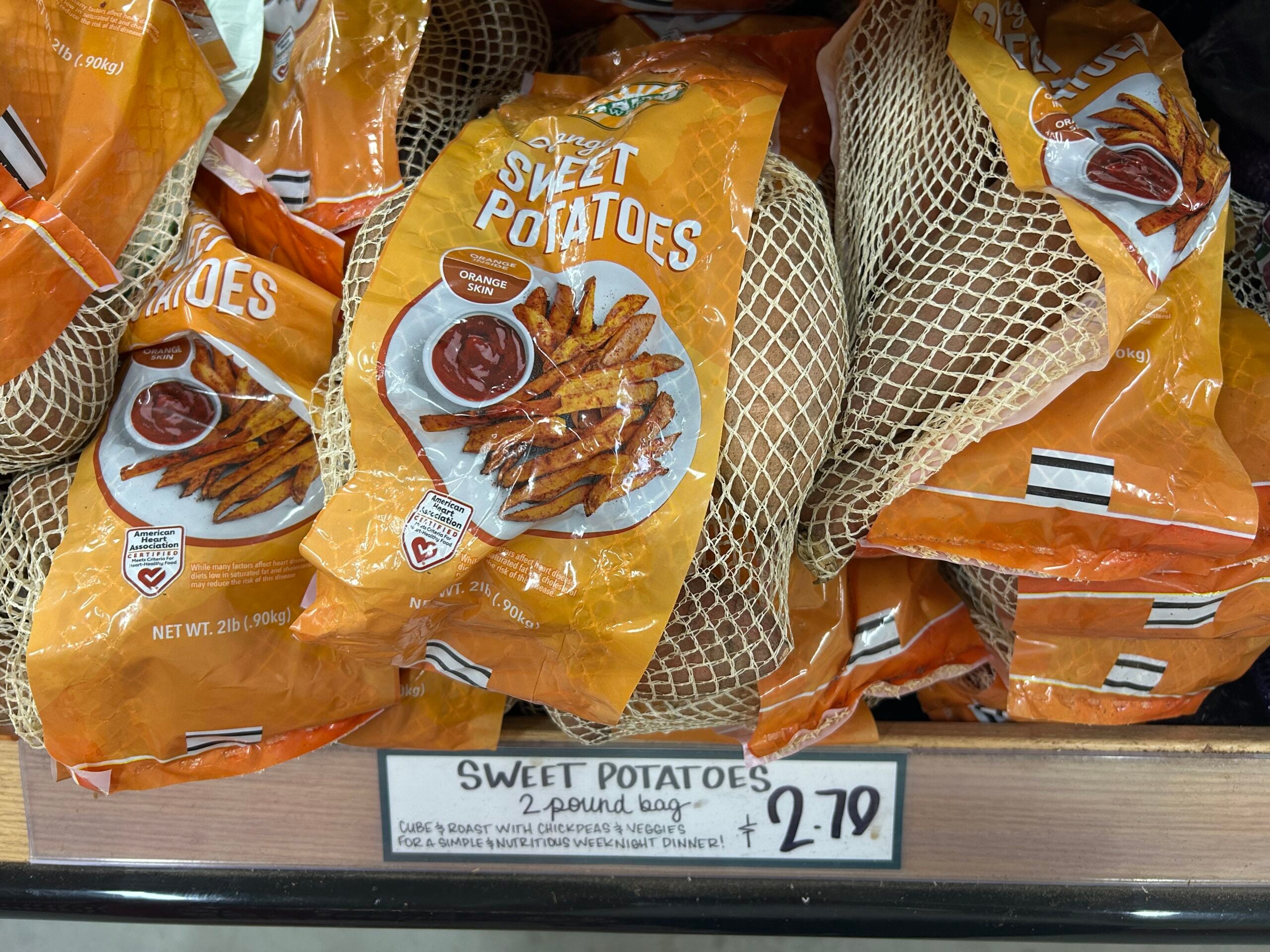 Bags of organic sweet potatoes in orange packaging are displayed on a grocery store shelf. A handwritten sign below reads “Sweet Potatoes 2 pound bag $2.79.”