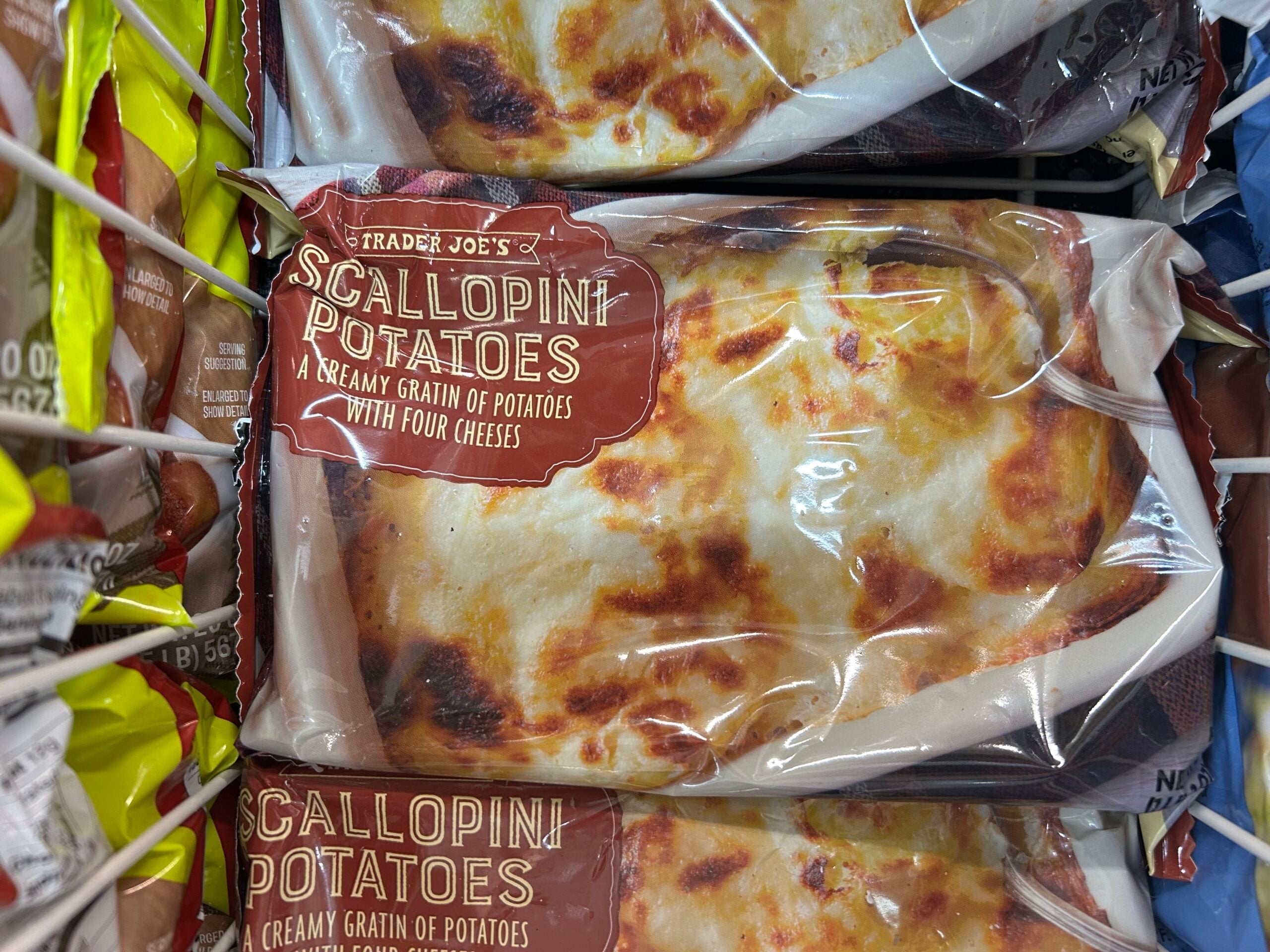 A frozen package of Trader Joe’s Scallopin’ Potatoes, described as a creamy gratin of potatoes with four cheeses, is shown on a grocery store shelf.