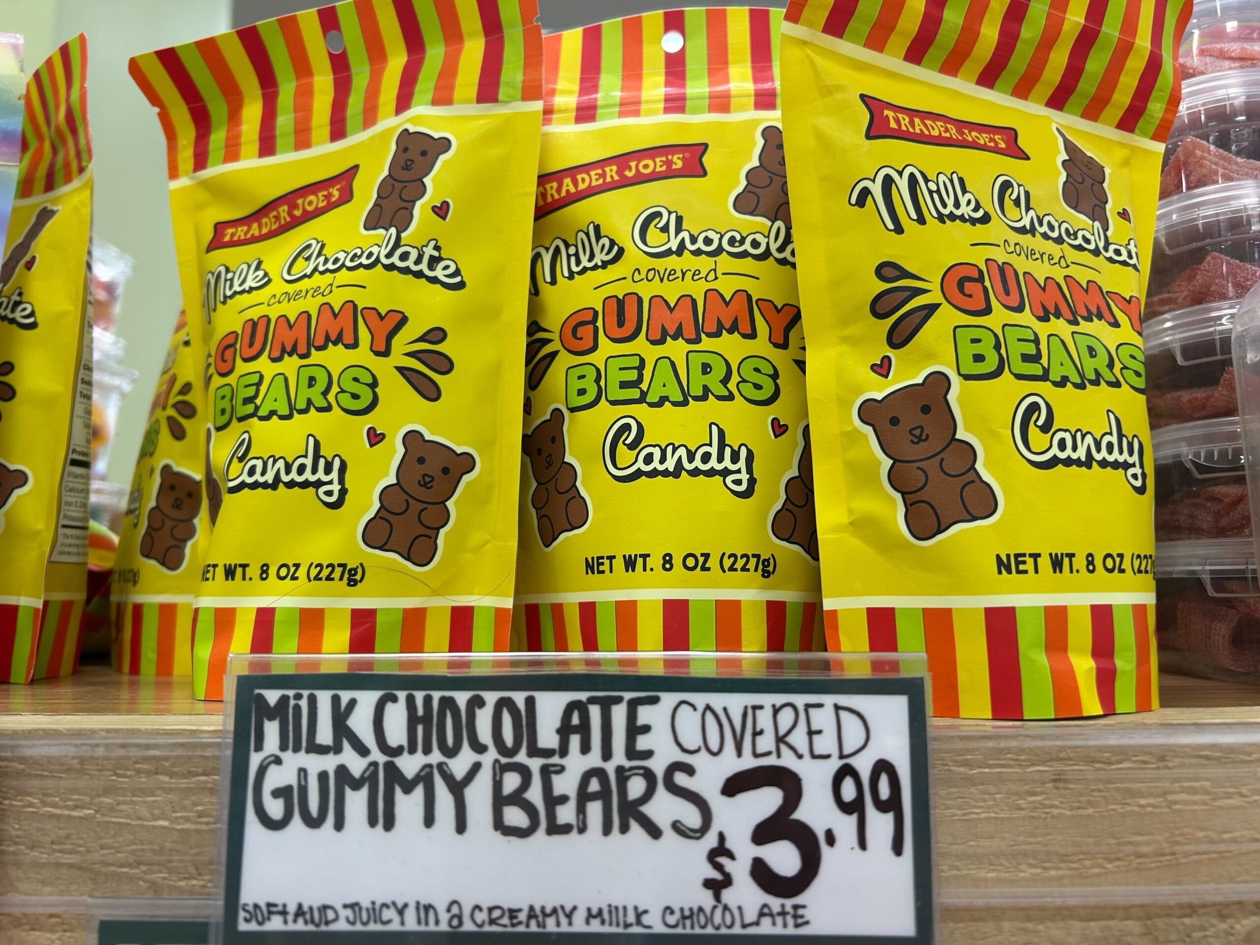 Bags of Trader Joe’s Milk Chocolate Covered Gummy Bears are displayed on a shelf above a handwritten price sign showing $3.99. The packaging is yellow with cartoon gummy bears and colorful lettering.