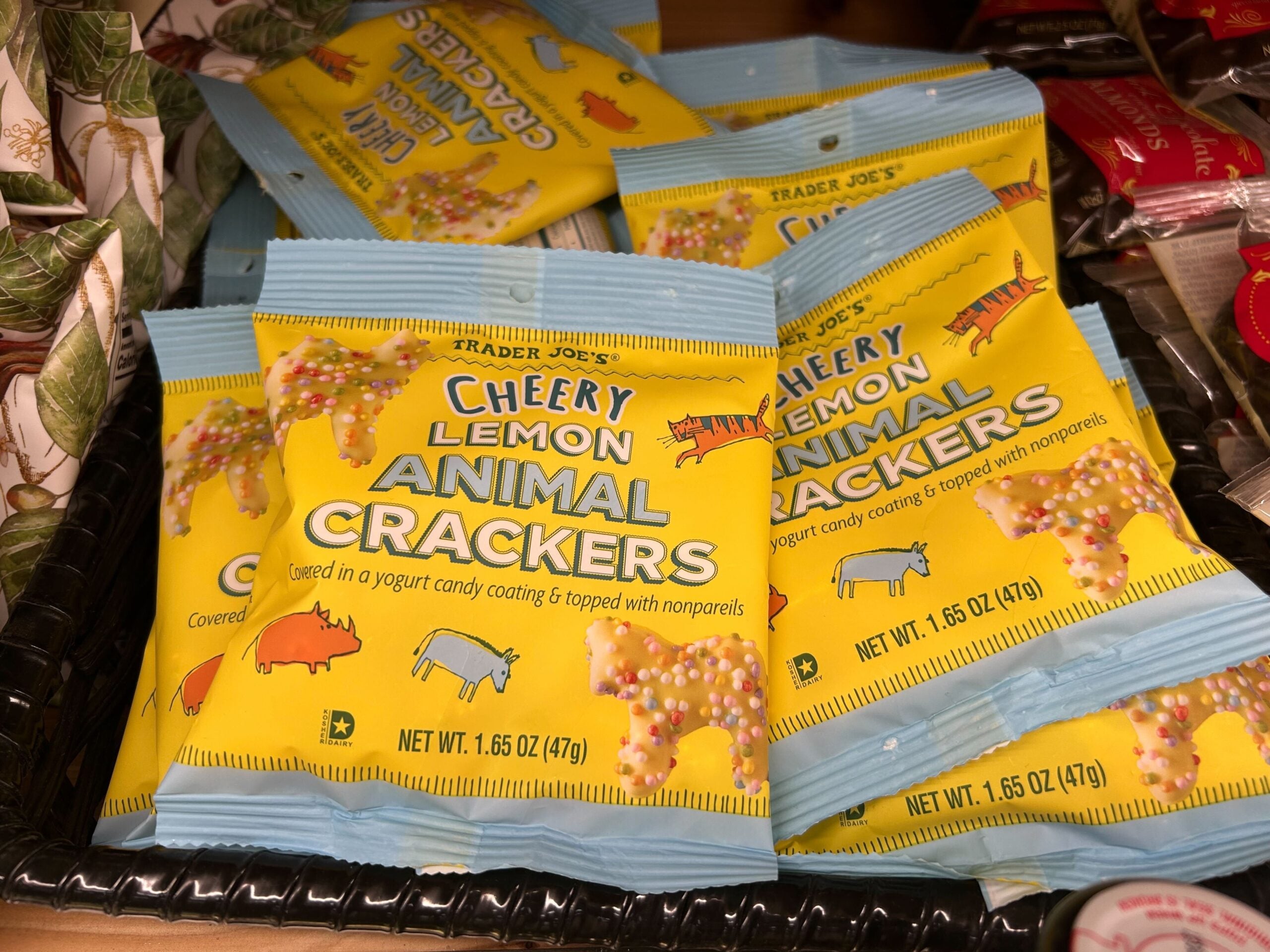 A display of Trader Joe's Cheery Lemon Animal Crackers in yellow and blue packaging, showing animal illustrations and labeled as covered in yogurt candy coating with nonpareils. Each bag is 1.65 oz (47g).
