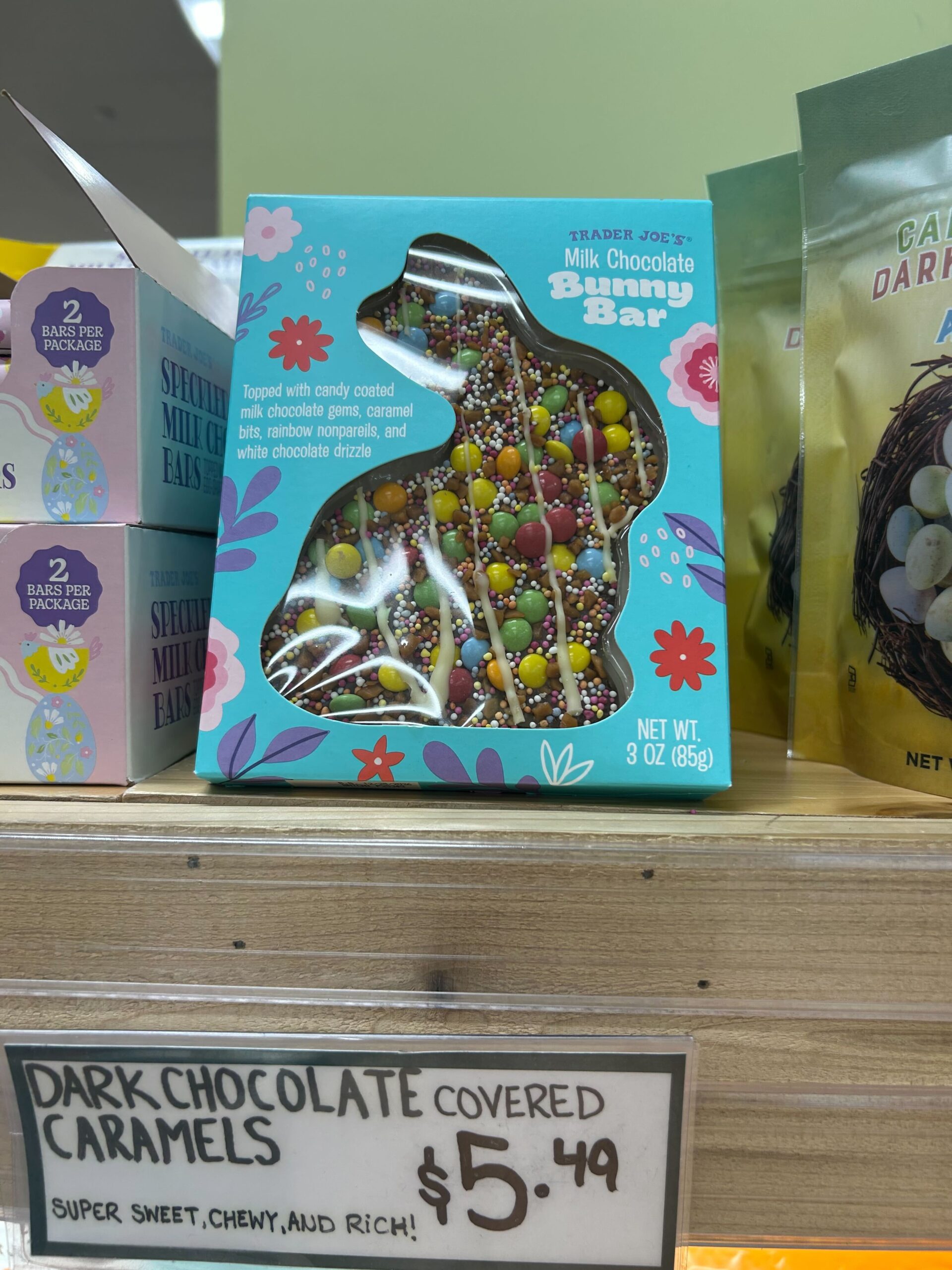 A bunny-shaped milk chocolate bar with colorful candy and sprinkles in festive packaging sits on a wooden shelf at Trader Joe’s. A sign below advertises dark chocolate covered caramels for $5.49.