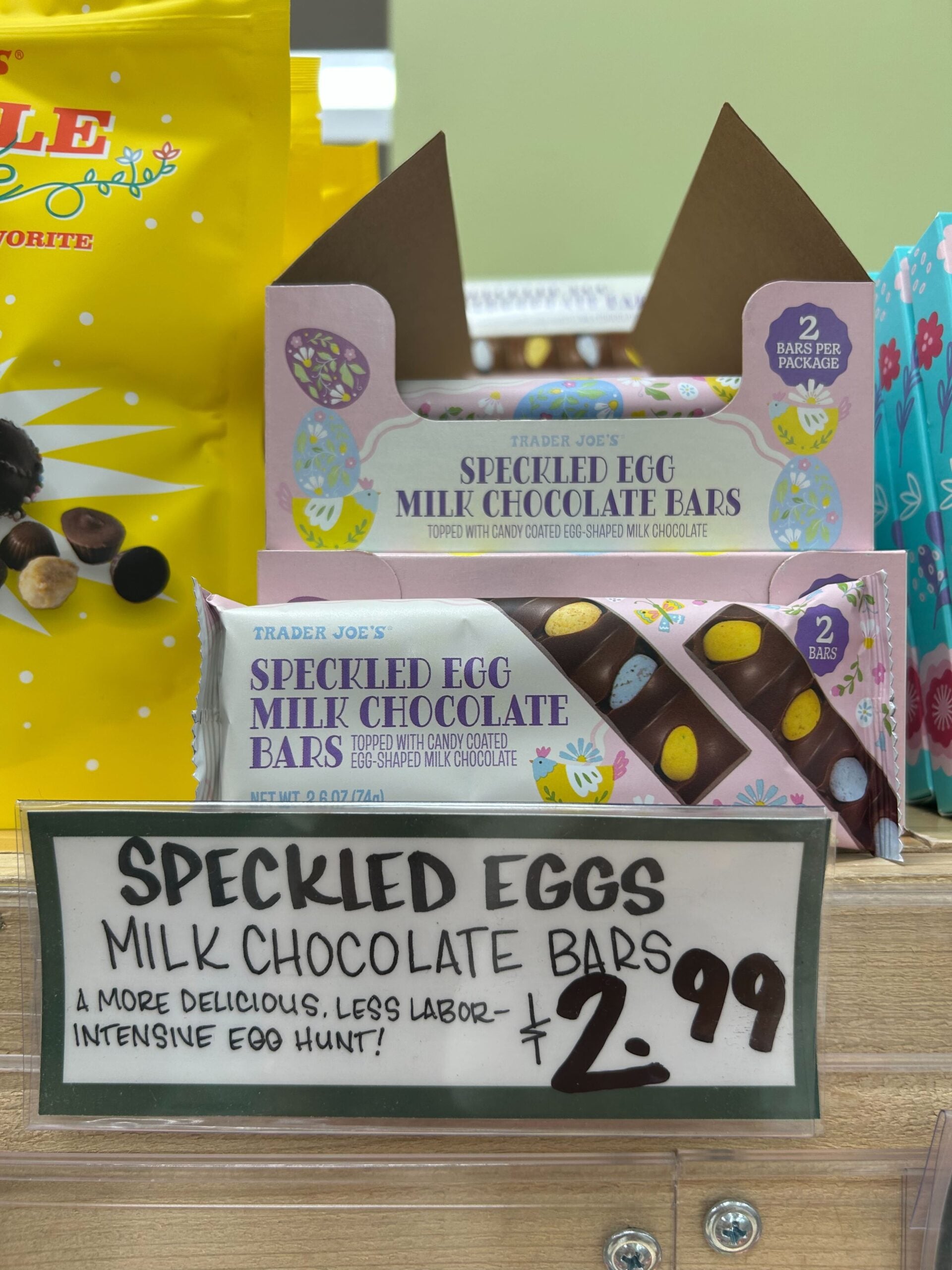 Trader Joe’s Speckled Egg Milk Chocolate Bars are displayed in an open box, with a sign below reading “SPECKLED EGGS MILK CHOCOLATE BARS, $2.99” and describing them as a less labor-intensive egg hunt.